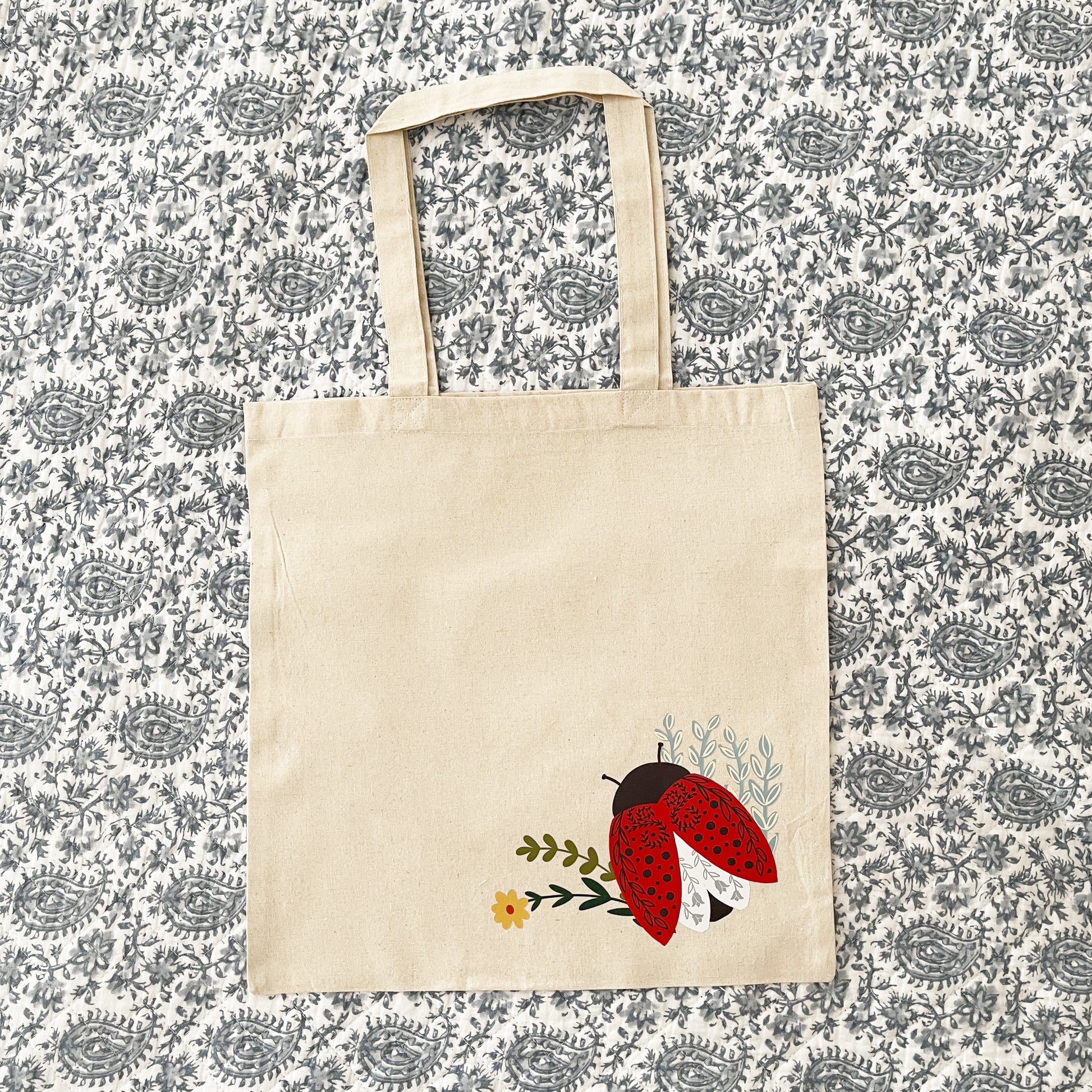 Beige canvas tote bag featuring a colorful illustration of a red ladybug with black spots, delicate floral accents, and green leaves near the bottom corner on a paisley background.