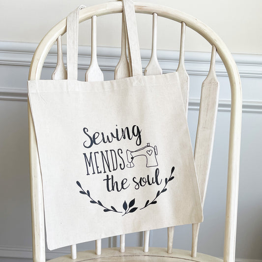 Natural cotton tote bag hanging on a white wooden chair, featuring the phrase 'Sewing mends the soul' in black script with a small illustration of a sewing machine and decorative leafy branches below. The tote has a minimalist, crafty design, perfect for sewing enthusiasts.