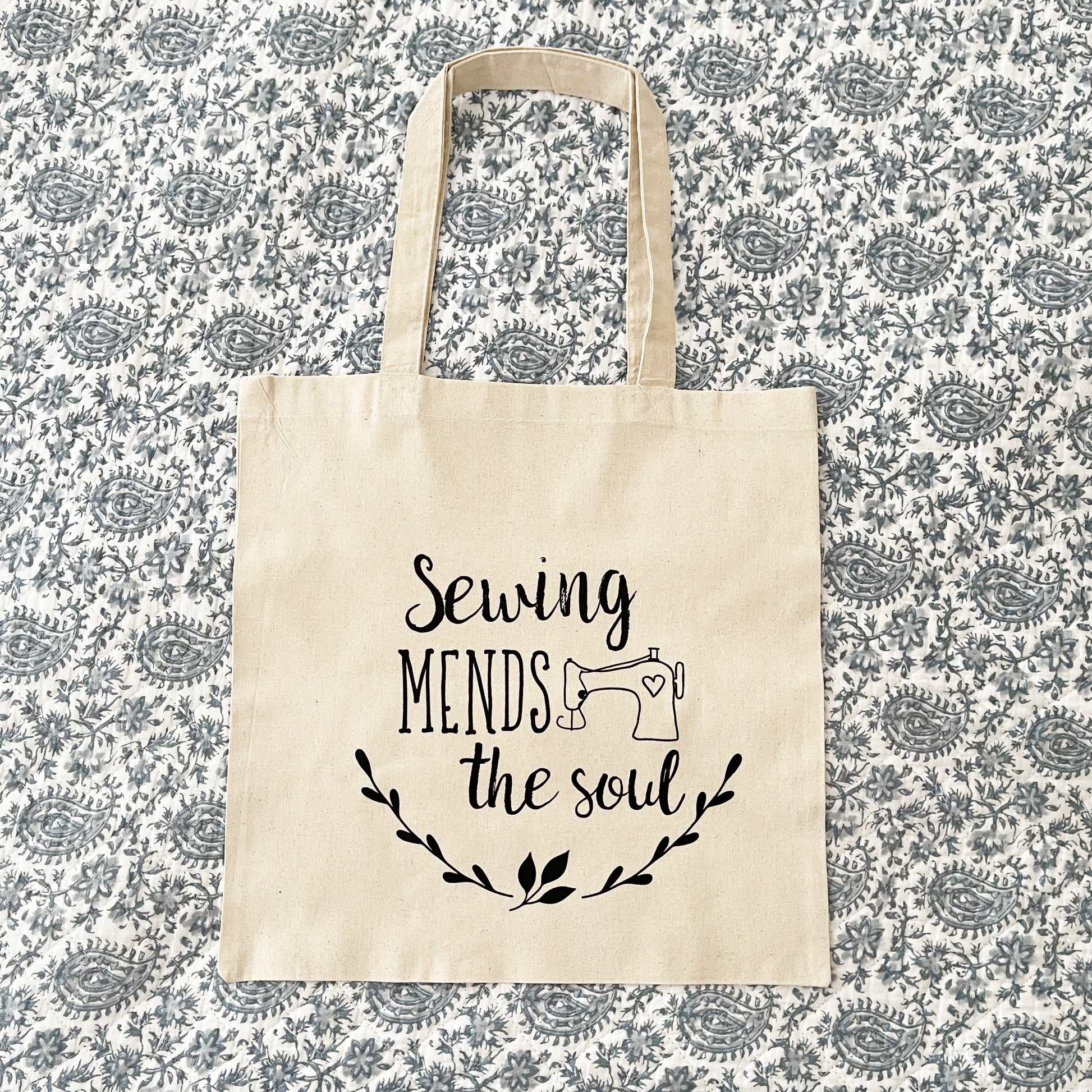 Natural cotton tote bag laying on a paisley background, featuring the phrase 'Sewing mends the soul' in black script with a small illustration of a sewing machine and decorative leafy branches below.