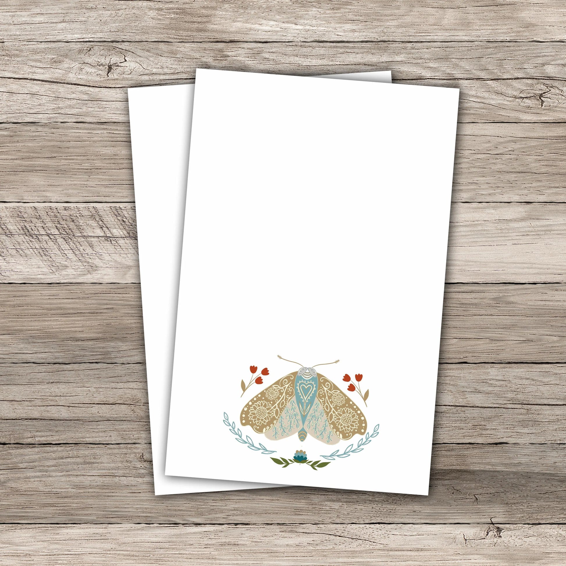 Two white notepads featuring an illustration of a moth with  mandala-like pattern wings  in soft beige and light blue, surrounded by small red tulip flowers and leafy green branches. The notepad rests on a rustic wooden background.