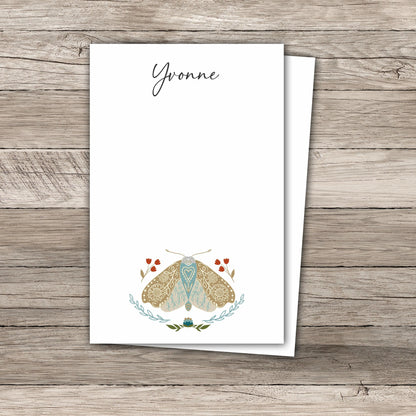 Two white notepads featuring an illustration of a moth with  mandala-like pattern wings  in soft beige and light blue, surrounded by small red tulip flowers and leafy green branches. The notepad rests on a rustic wooden background.