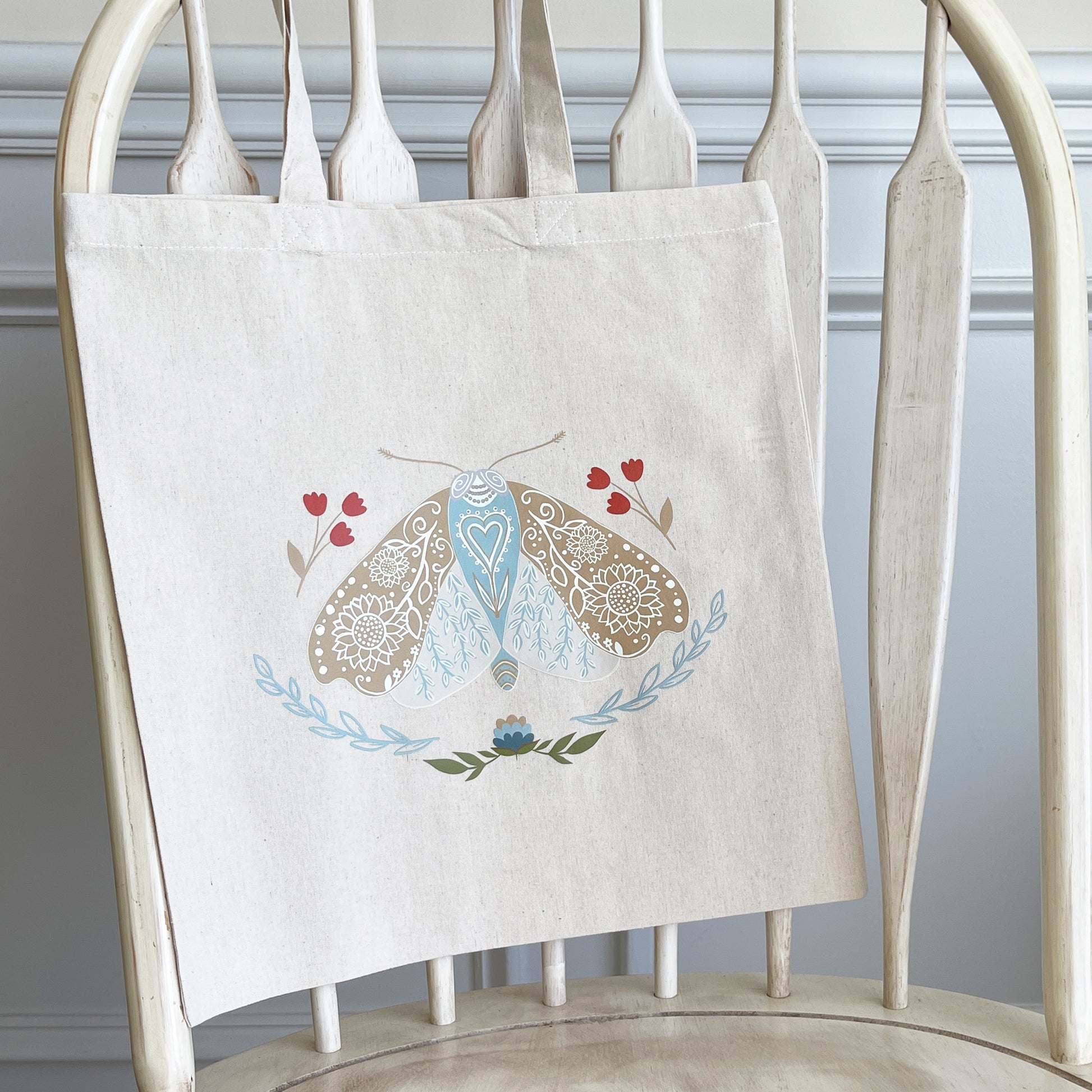 Canvas tote bag hanging on a white wooden chair featuring an illustration of a detailed moth with blue, beige, and red floral elements surrounding it.