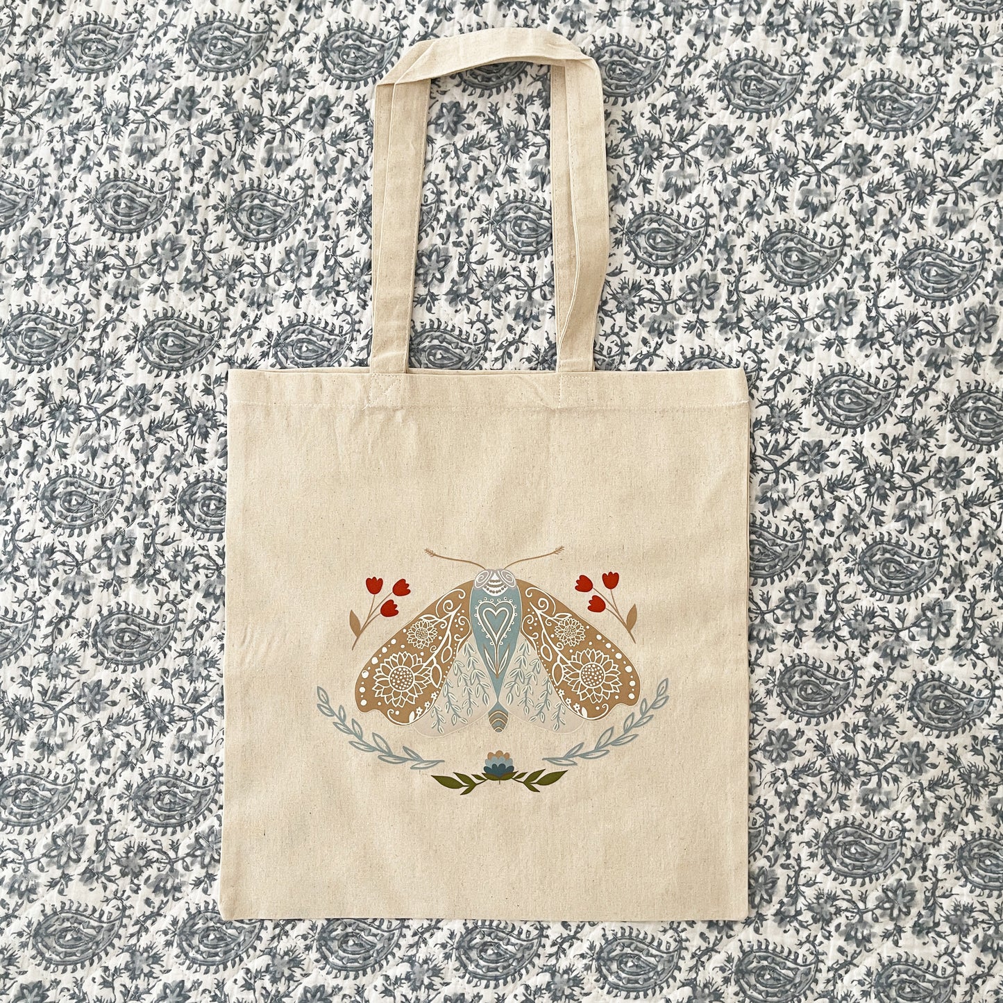 Canvas tote bag featuring an illustration of a detailed moth with blue, beige, and red floral elements surrounding it.