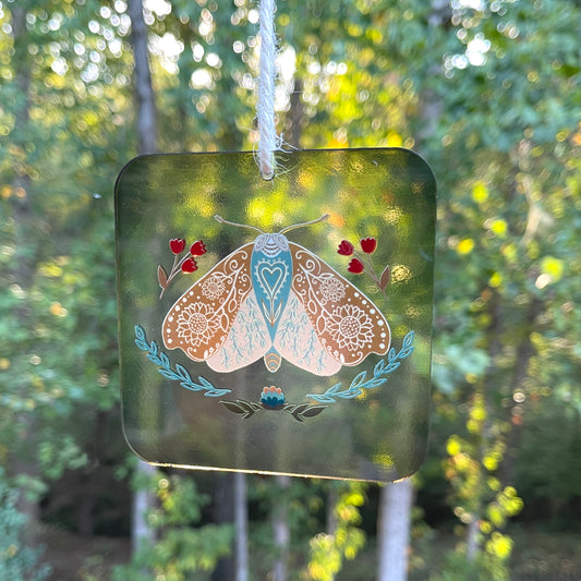 Transparent acrylic suncatcher featuring a illustration of an intricate moth with beige wings adorned with floral patterns, framed by small red tulips and leafy branches, hanging by a ribbon with a blurred tree background.