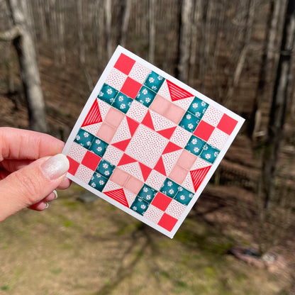 A colorful quilt pattern featuring a combination of teal, red, and peach squares and triangles arranged in a geometric design, held against a wooded background.