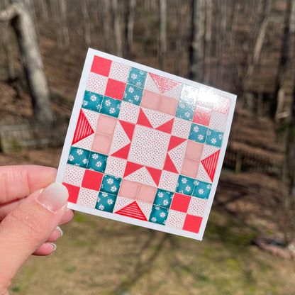 A colorful quilt pattern featuring a combination of teal, red, and peach squares and triangles arranged in a geometric design, held against a wooded background.