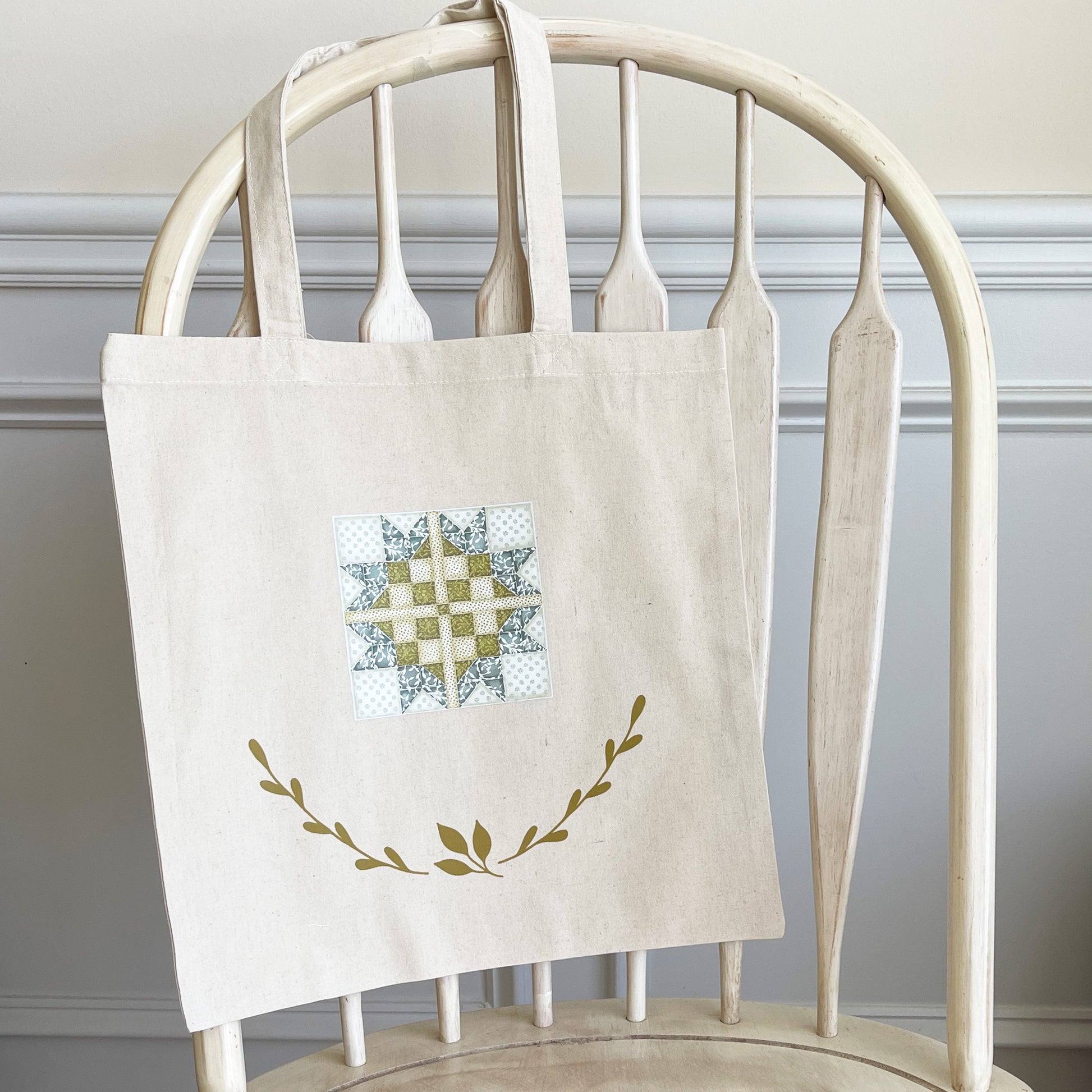 Natural cotton tote bag hanging on a white wooden chair, featuring a quilt block design in soft blue, green, and white tones. Below the quilt block, two green leafy branches curve upward, adding a decorative touch. 