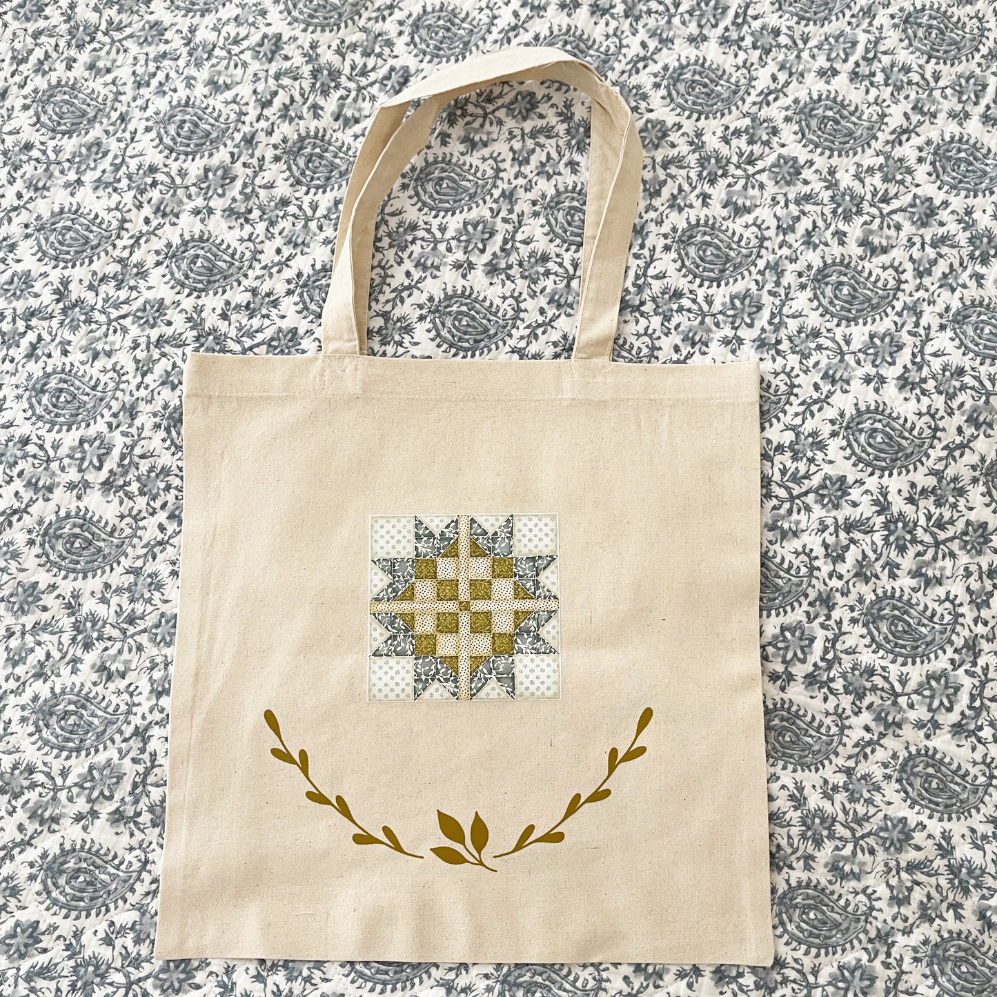 Natural cotton tote bag laying flat on a paisley background, featuring a quilt block design in soft blue, green, and white tones. Below the quilt block, two green leafy branches curve upward, adding a decorative touch.