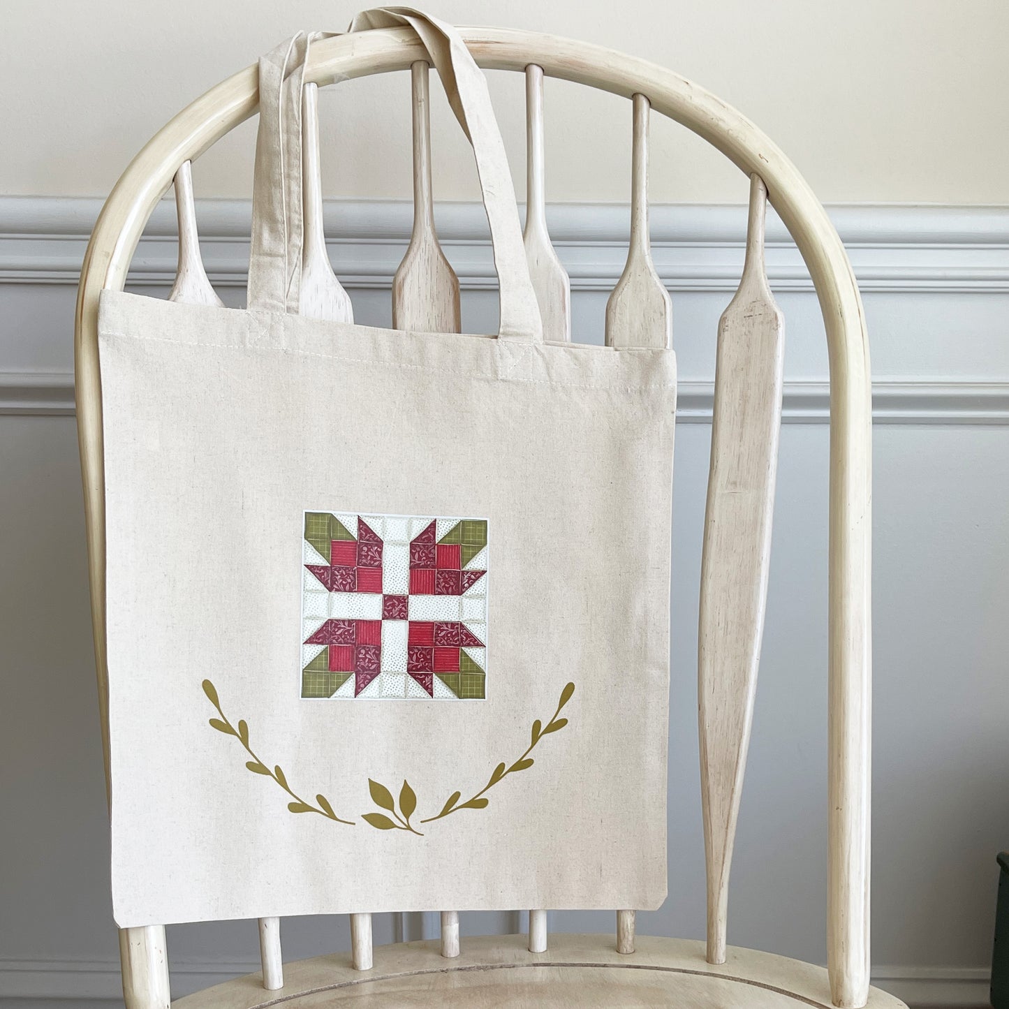 Natural cotton tote bag hanging on a white wooden chair, featuring a red and green quilt block design in the center. Below the quilt block, two green leafy branches curve upward, adding a decorative element.