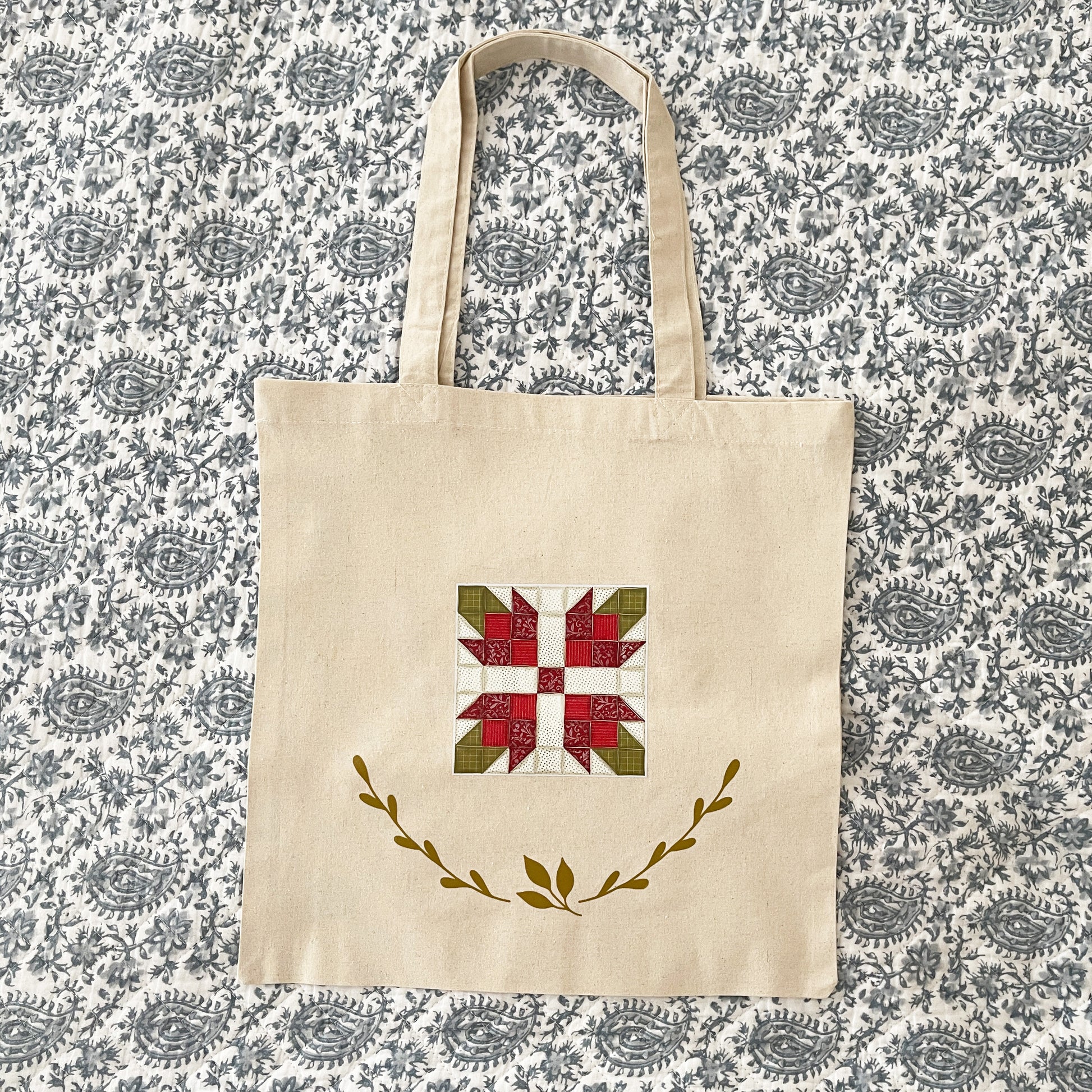 Natural cotton tote bag laying on a paisley background featuring a red and green quilt block design in the center. Below the quilt block, two green leafy branches curve upward, adding a decorative element.