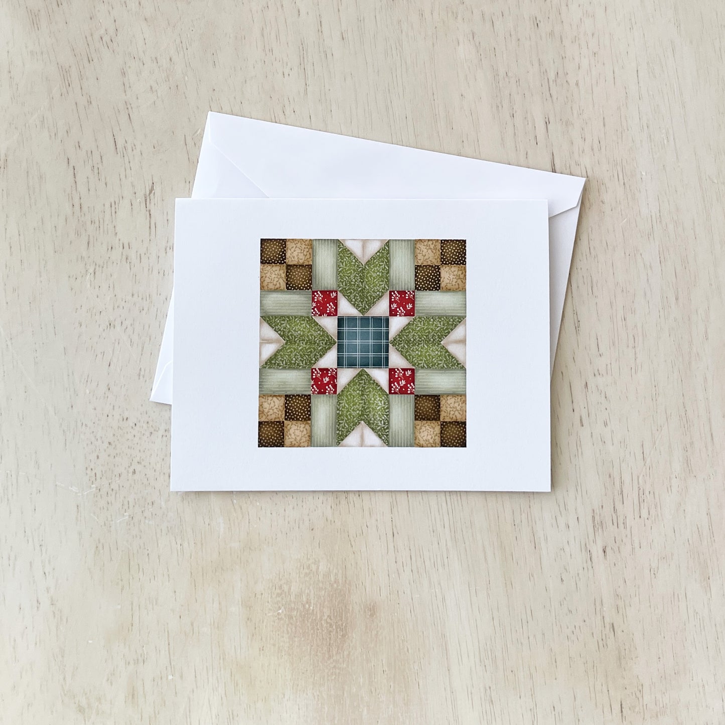 Handmade greeting card with a quilt pattern design featuring green, red, and beige fabric squares arranged in a star-like pattern, placed on a light wood surface with a white envelope beside it.