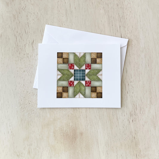 Handmade greeting card with a quilt pattern design featuring green, red, and beige fabric squares arranged in a star-like pattern, placed on a light wood surface with a white envelope beside it.