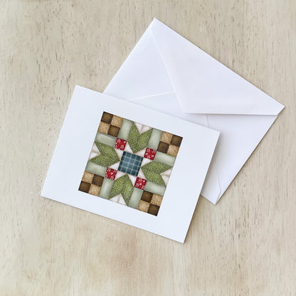 Handmade greeting card with a quilt pattern design featuring green, red, and beige fabric squares arranged in a star-like pattern, placed on a light wood surface with a white envelope beside it.