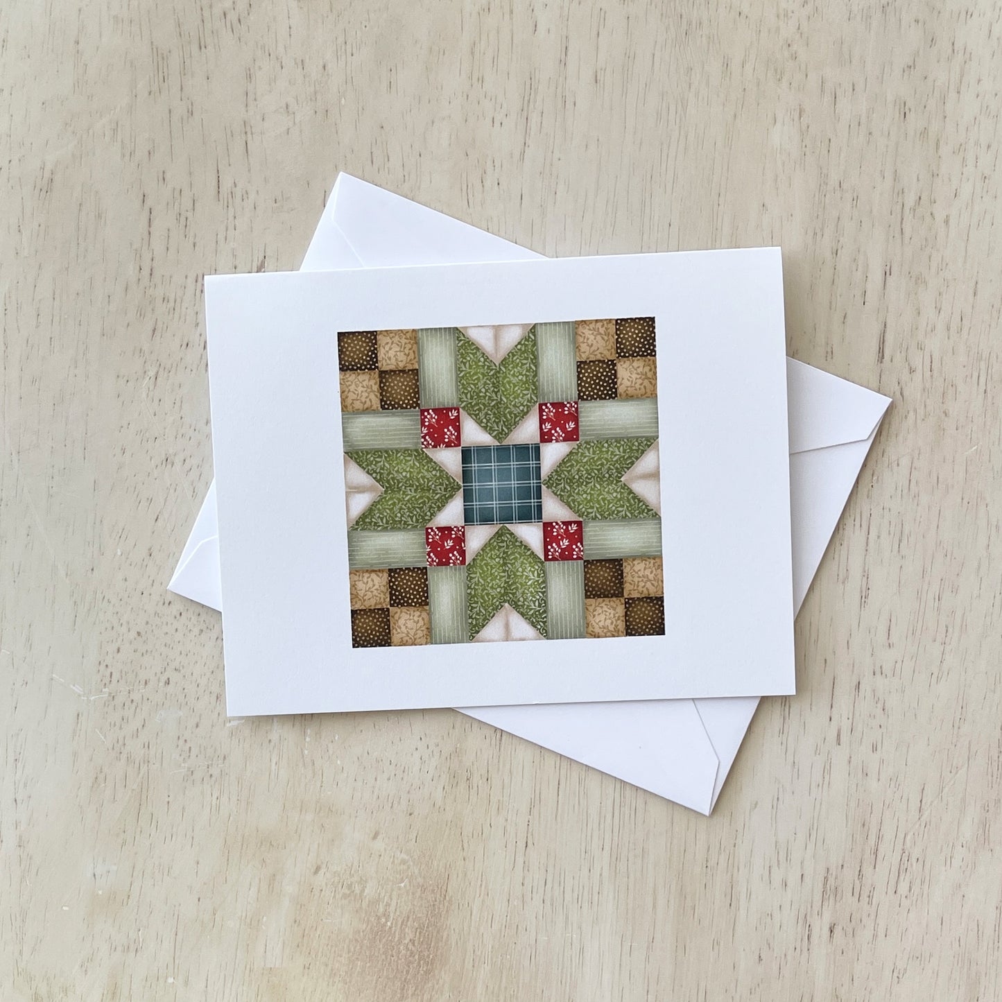 Handmade greeting card with a quilt pattern design featuring green, red, and beige fabric squares arranged in a star-like pattern, placed on a light wood surface with a white envelope beside it.