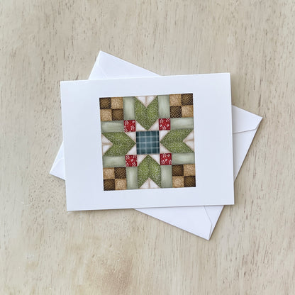 Handmade greeting card with a quilt pattern design featuring green, red, and beige fabric squares arranged in a star-like pattern, placed on a light wood surface with a white envelope beside it.