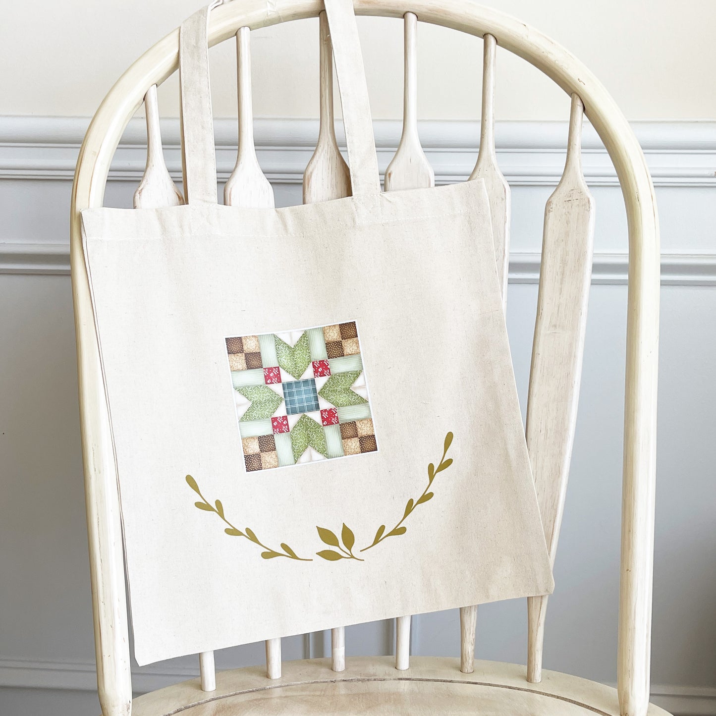 Natural cotton tote bag hanging on a white wooden chair, featuring a colorful quilt block design in the center with red, green, and beige patterns. Below the quilt block, two green leafy branches curve upward, adding a decorative touch.