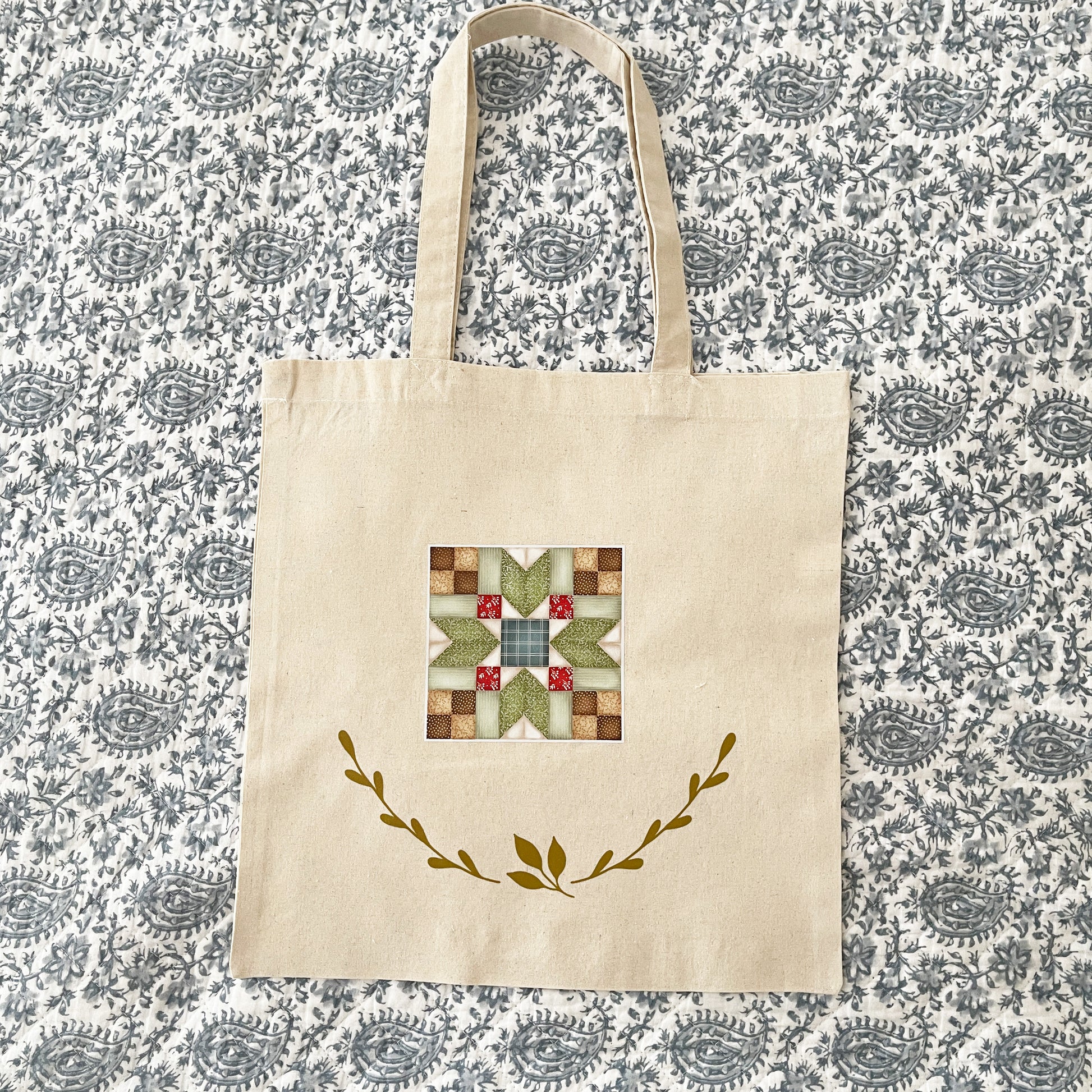 Natural cotton tote bag laying on a paisley background, featuring a colorful quilt block design in the center with red, green, and beige patterns. Below the quilt block, two green leafy branches curve upward, adding a decorative touch.