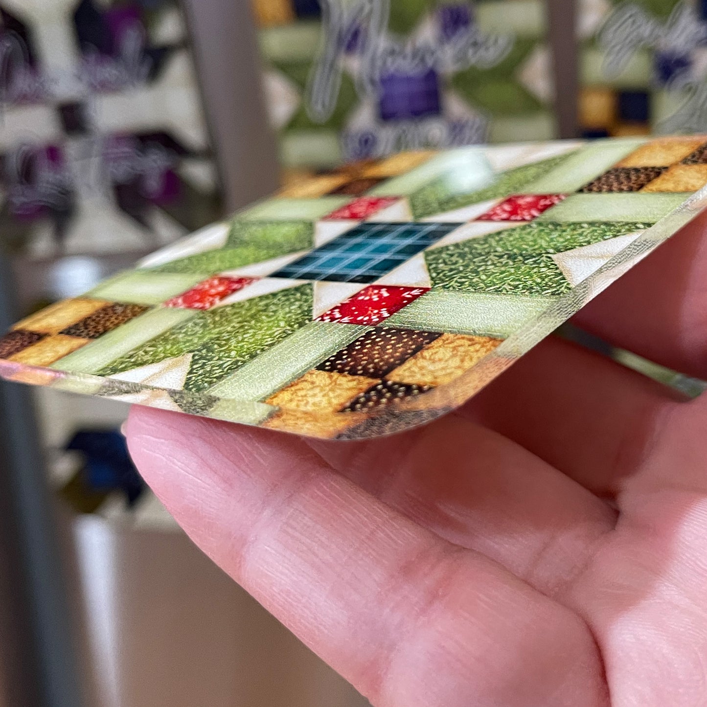 A close-up side view of a hand holding a thin, square magnet with a quilt pattern design featuring green, red, and beige colors. The background shows additional magnets with similar quilt patterns and personalized text.