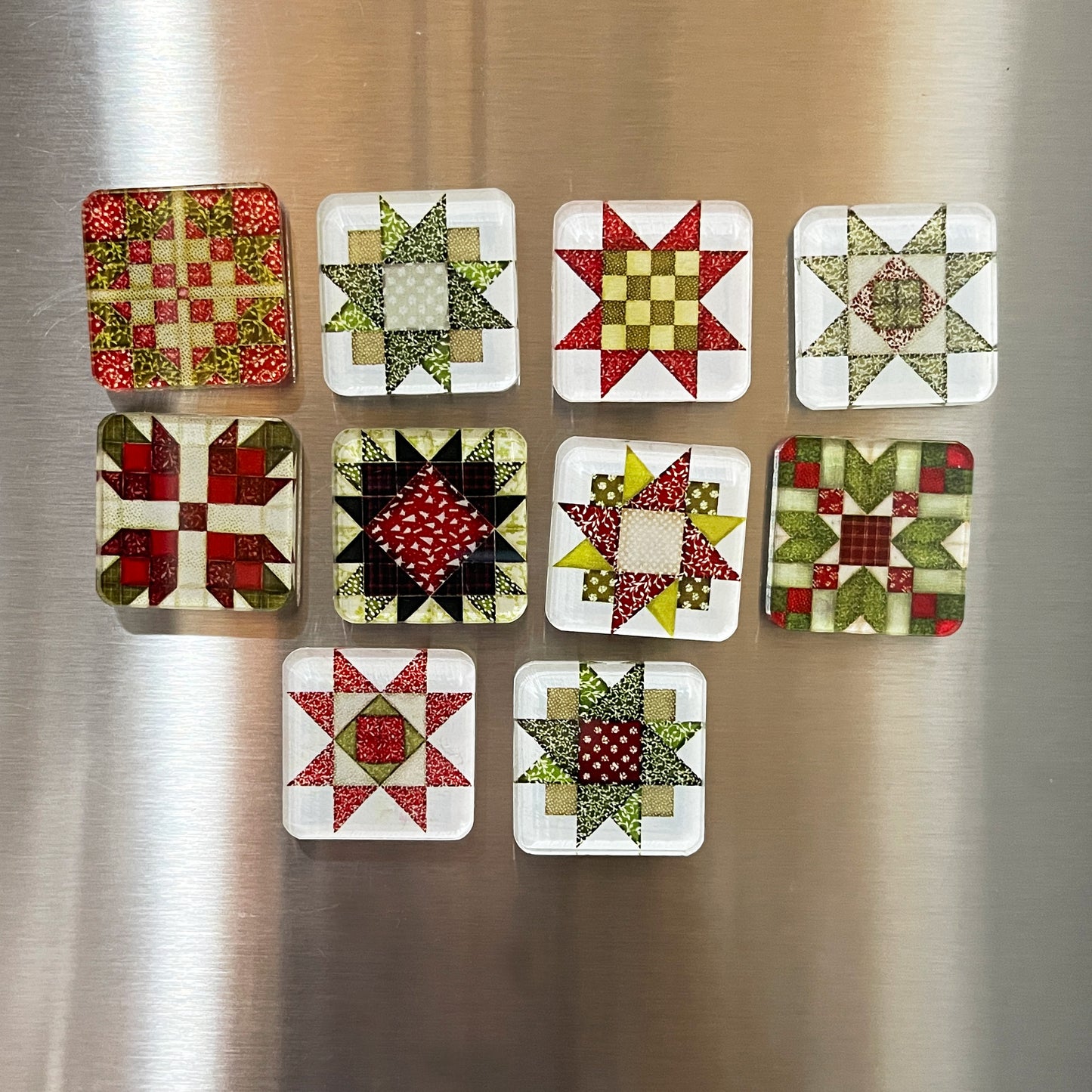 Set of ten square magnets featuring colorful Christmas-themed quilt block designs in traditional red, green, and white patterns. Each magnet showcases a unique geometric patchwork design, arranged in a neat grid on a metallic background.