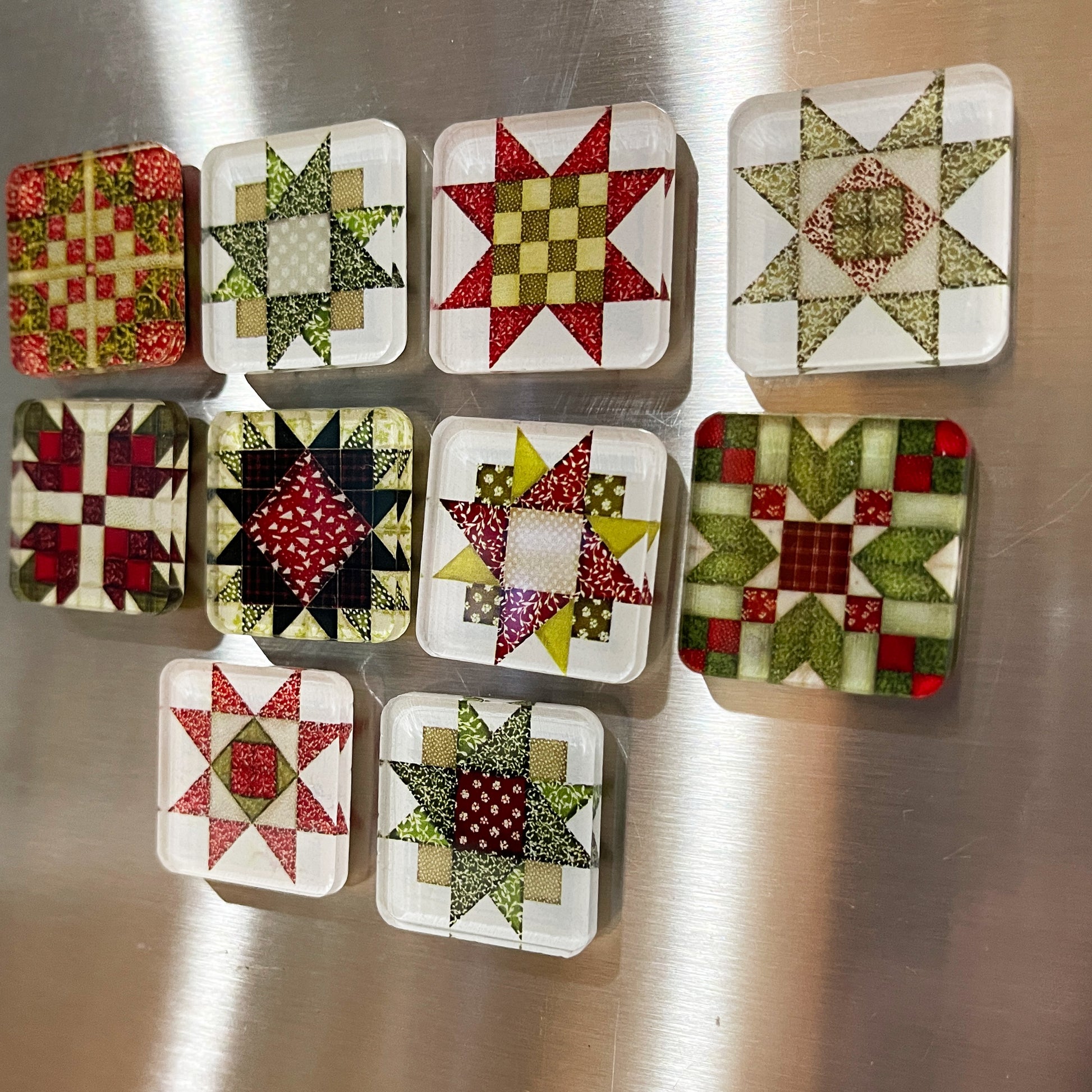 Set of ten square magnets featuring colorful Christmas-themed quilt block designs in traditional red, green, and white patterns. Each magnet showcases a unique quilt patchwork design, arranged in a neat grid on a metallic background.