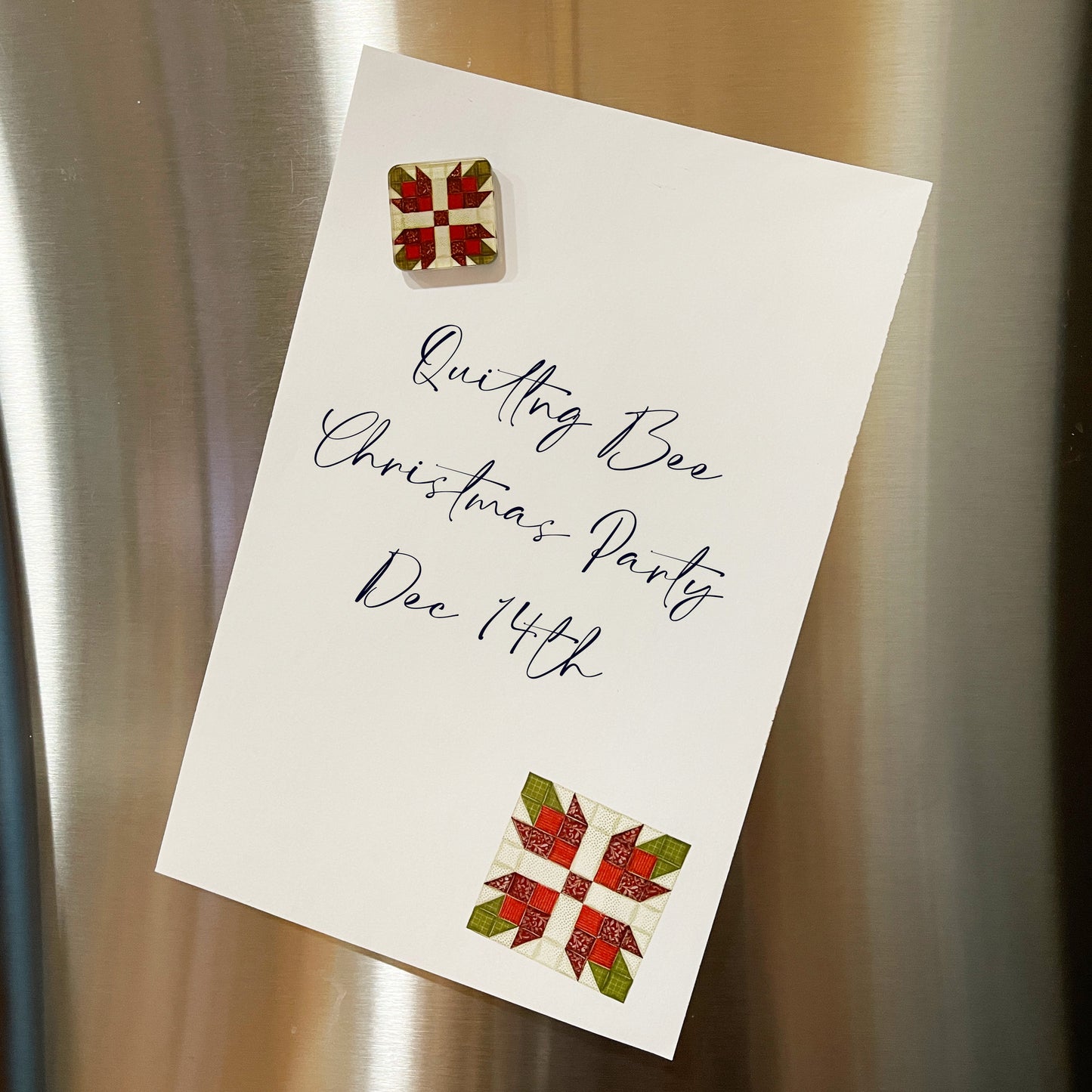 Christmas-themed quilt block magnet holding a note on a stainless steel refrigerator. The magnet features a red and green geometric patchwork design, securing a handwritten invitation that reads 'Quilting Bee Christmas Party Dec 14th' in elegant script.