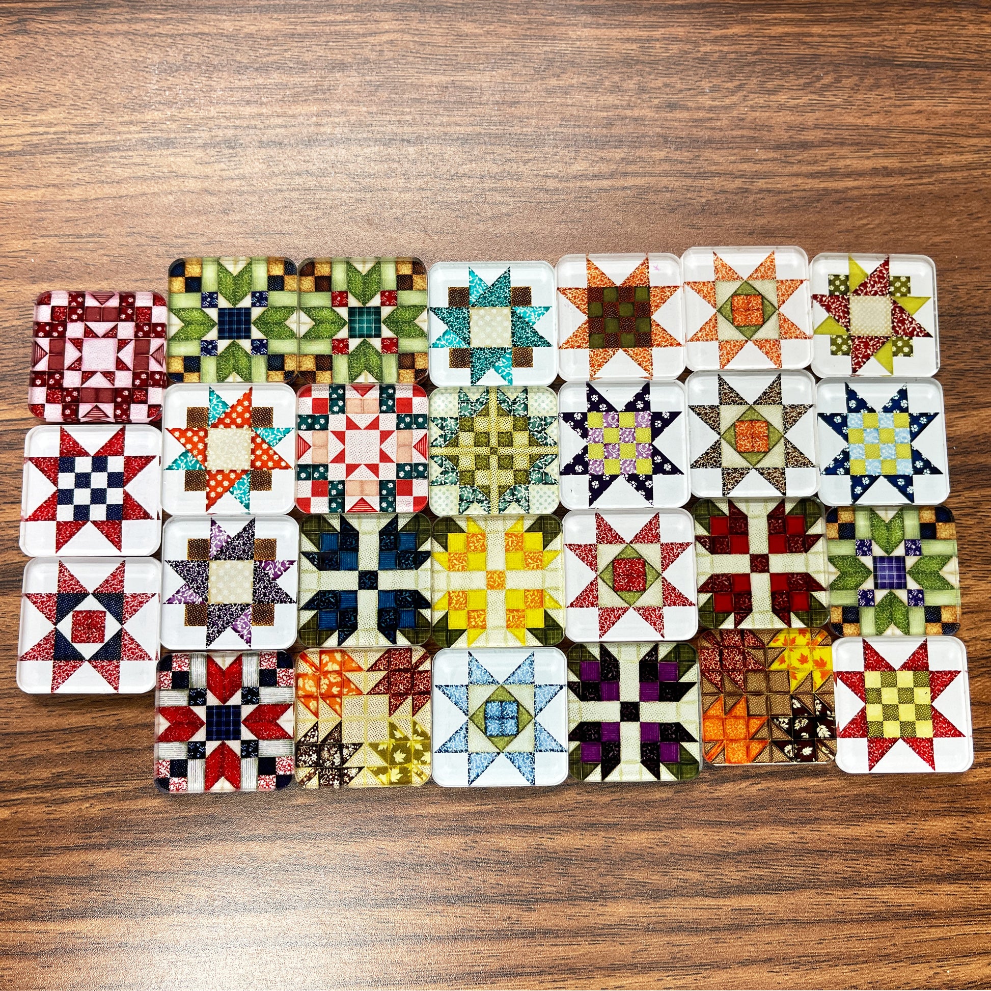 Square magnets featuring various quilt block designs in different colors, including red, green, blue, and yellow. The magnets are arranged on a light wooden surface, each showcasing a unique patchwork pattern.