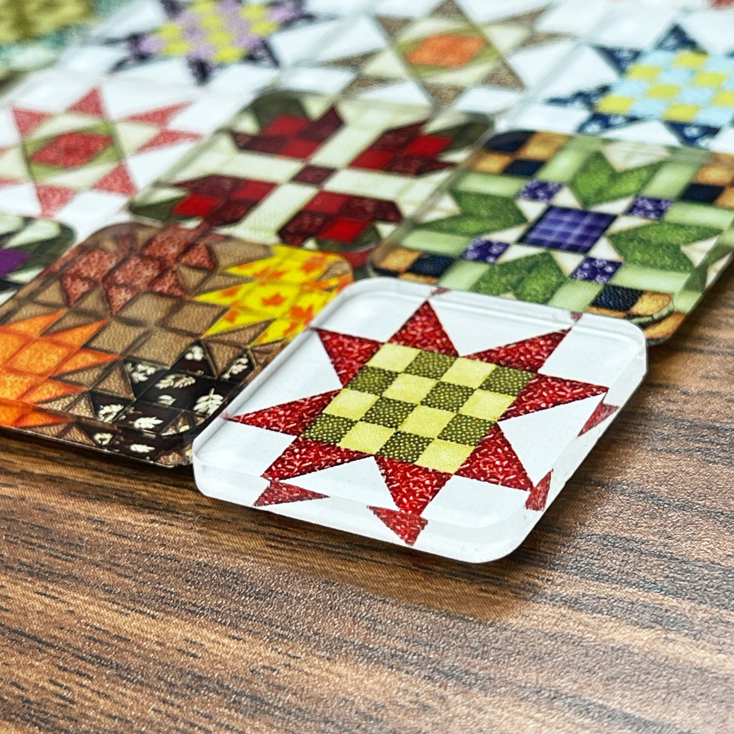 Close up side view square magnets featuring various quilt block designs in different colors, including red, green, blue, and yellow. The magnets are arranged on a dark wooden surface, each showcasing a unique patchwork pattern.