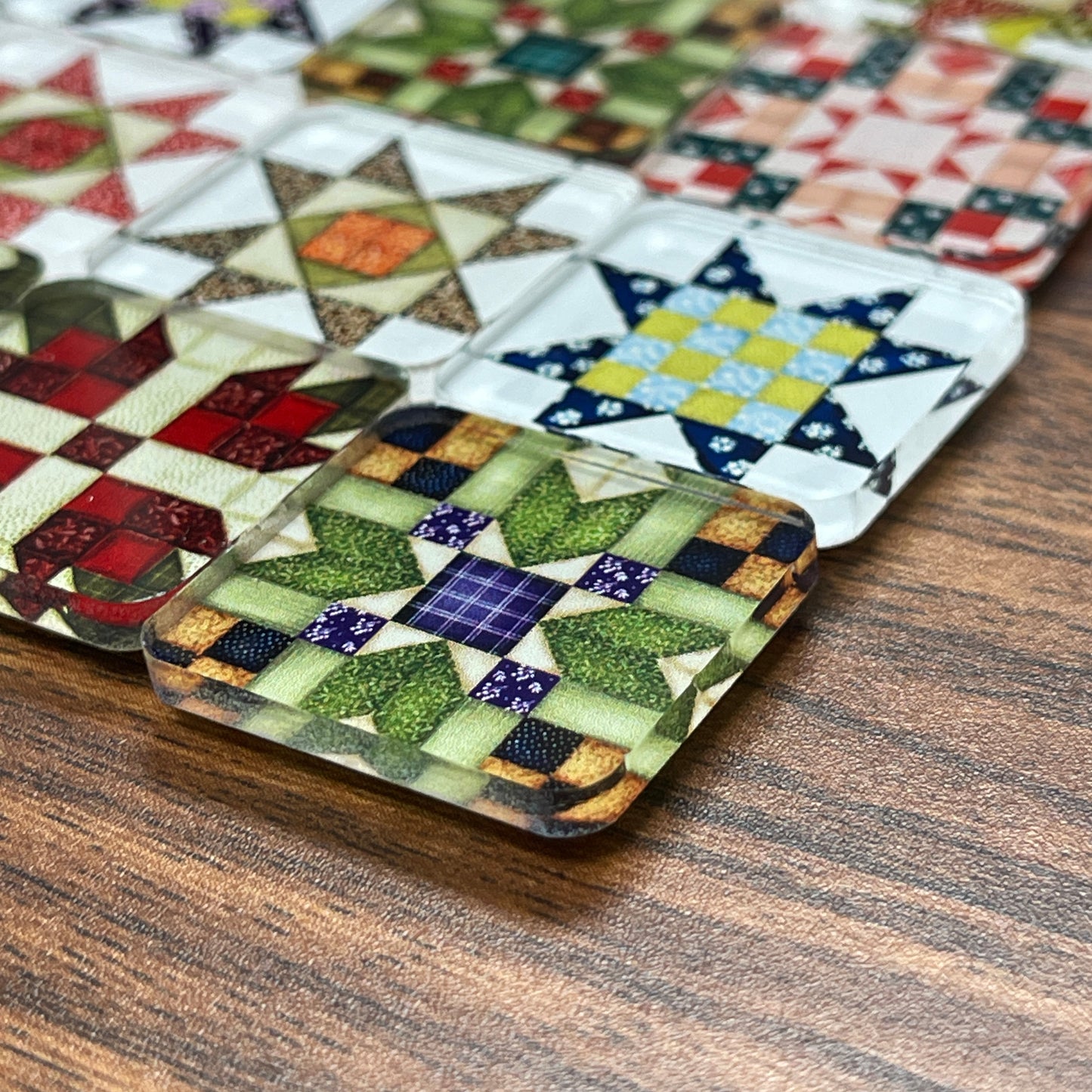 Close up side view square magnets featuring various quilt block designs in different colors, including red, green, blue, and yellow. The magnets are arranged on a dark wooden surface, each showcasing a unique patchwork pattern.