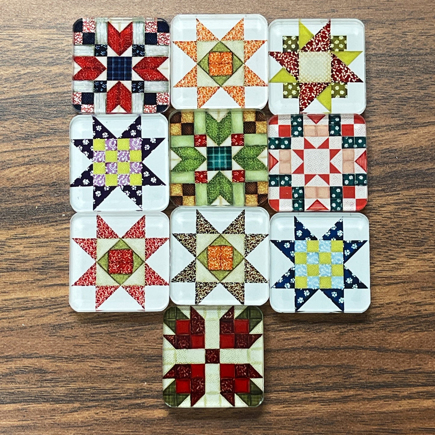 Set of ten square magnets featuring various quilt block designs in different colors, including red, green, blue, and yellow. The magnets are arranged on a dark wooden surface, each showcasing a unique patchwork pattern.