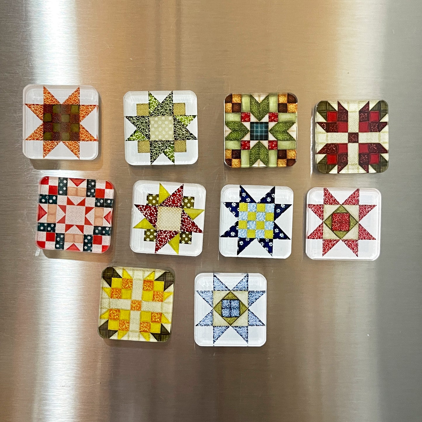 Set of ten square magnets featuring various quilt block designs in different colors, including red, green, blue, and yellow. The magnets are arranged on a stainless steel surface, each showcasing a unique patchwork pattern.