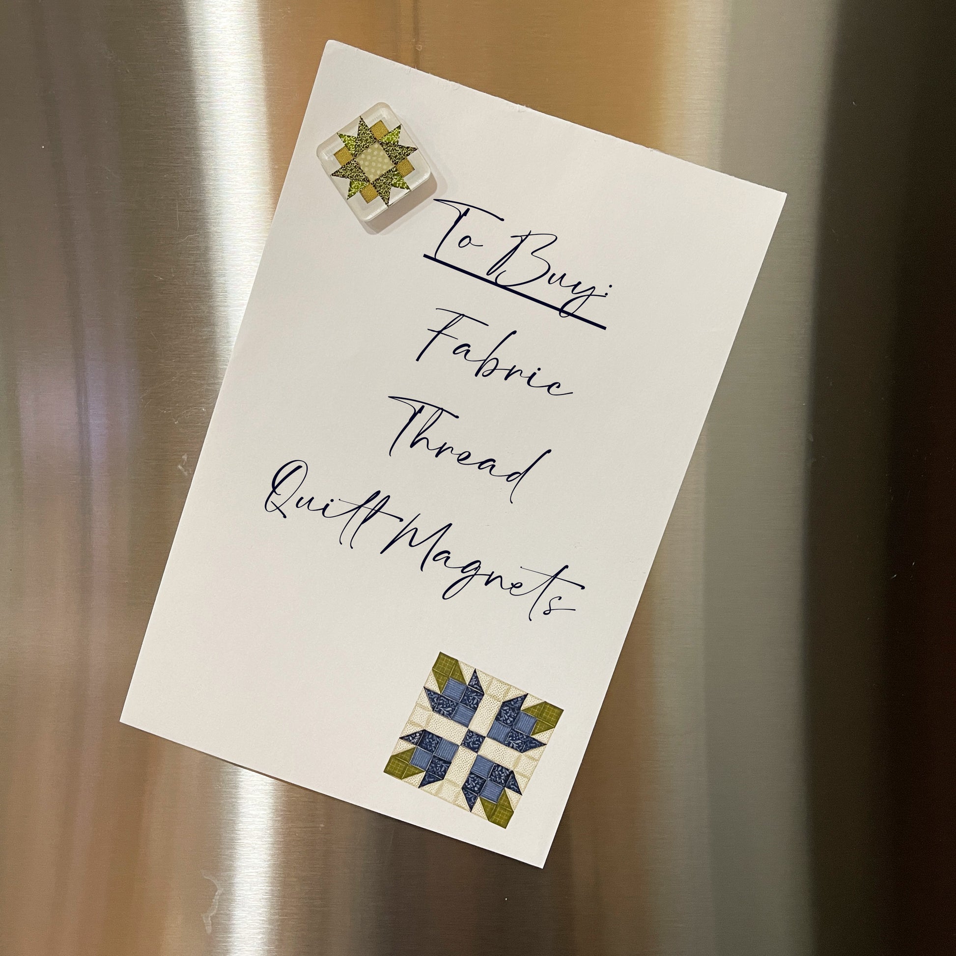 Green quilt block magnet holding a  note on a stainless steel refrigerator. The blue floral note has 'To Buy' list written in script.