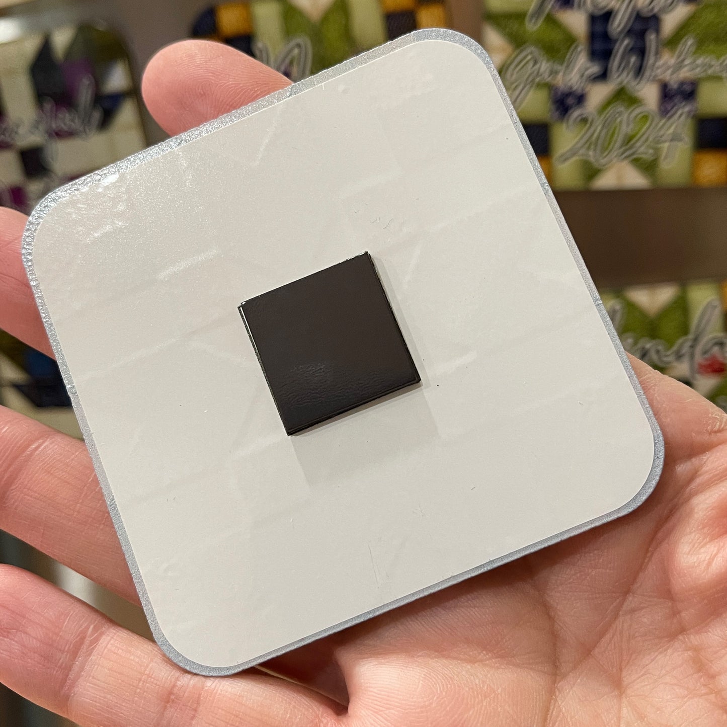 A close-up of the back of a square magnet held in a hand. The magnet has a white backing with a small black square magnet in the center. The background shows additional magnets with quilt patterns and personalized text.