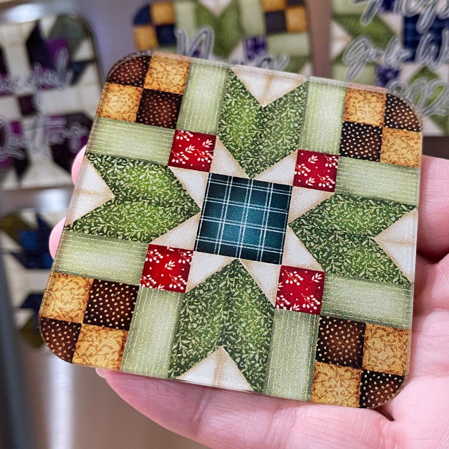 A close-up of a hand holding a square magnet featuring a quilt pattern with green, red, and beige colors. The background shows additional magnets with similar quilt designs and personalized text.