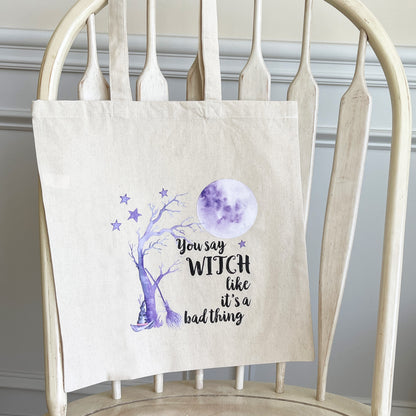 Beige canvas tote bag hanging on a wooden chair, featuring a whimsical purple design of a witch’s hat, a spooky tree, stars, and a full moon, with the text 'You say WITCH like it’s a bad thing' printed in black.