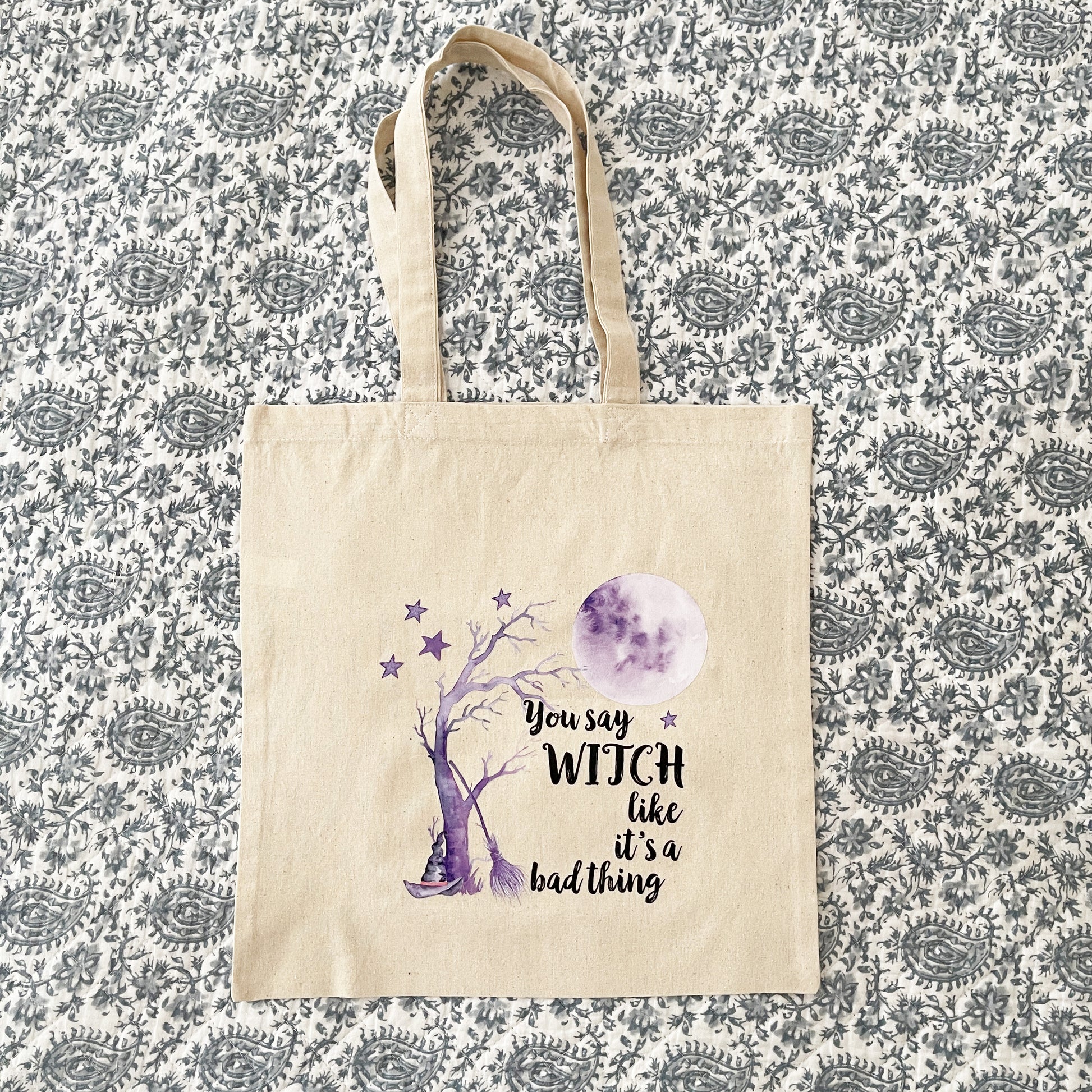 Beige canvas tote bag featuring a whimsical purple design of a witch’s hat, a spooky tree, stars, and a full moon, with the text 'You say WITCH like it’s a bad thing' printed in black.