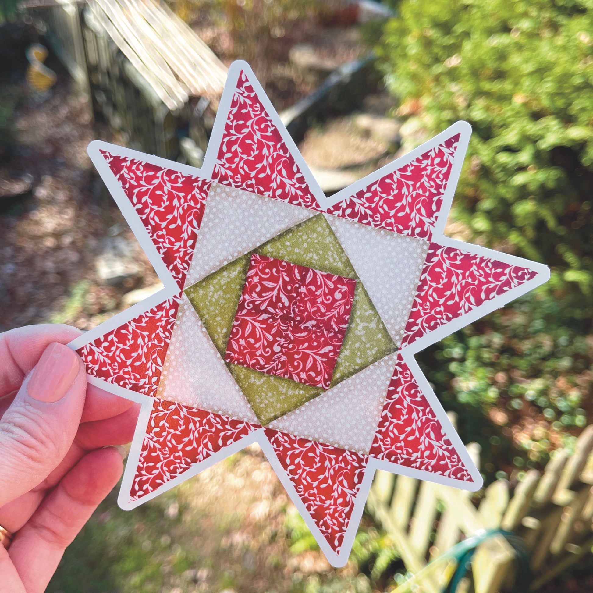 Large Red, Green and Tan Quilt block sticker held over grassy outside background. 