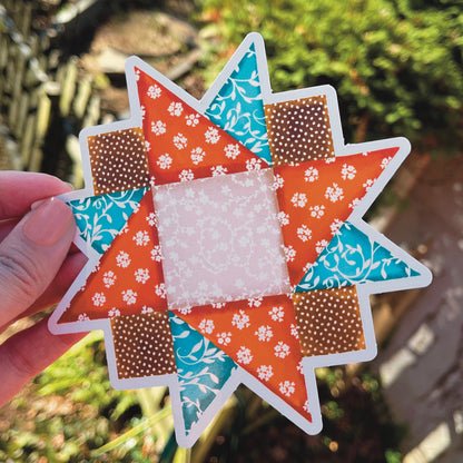 Large orange, blue and brown quilt block sticker held over grassy background.