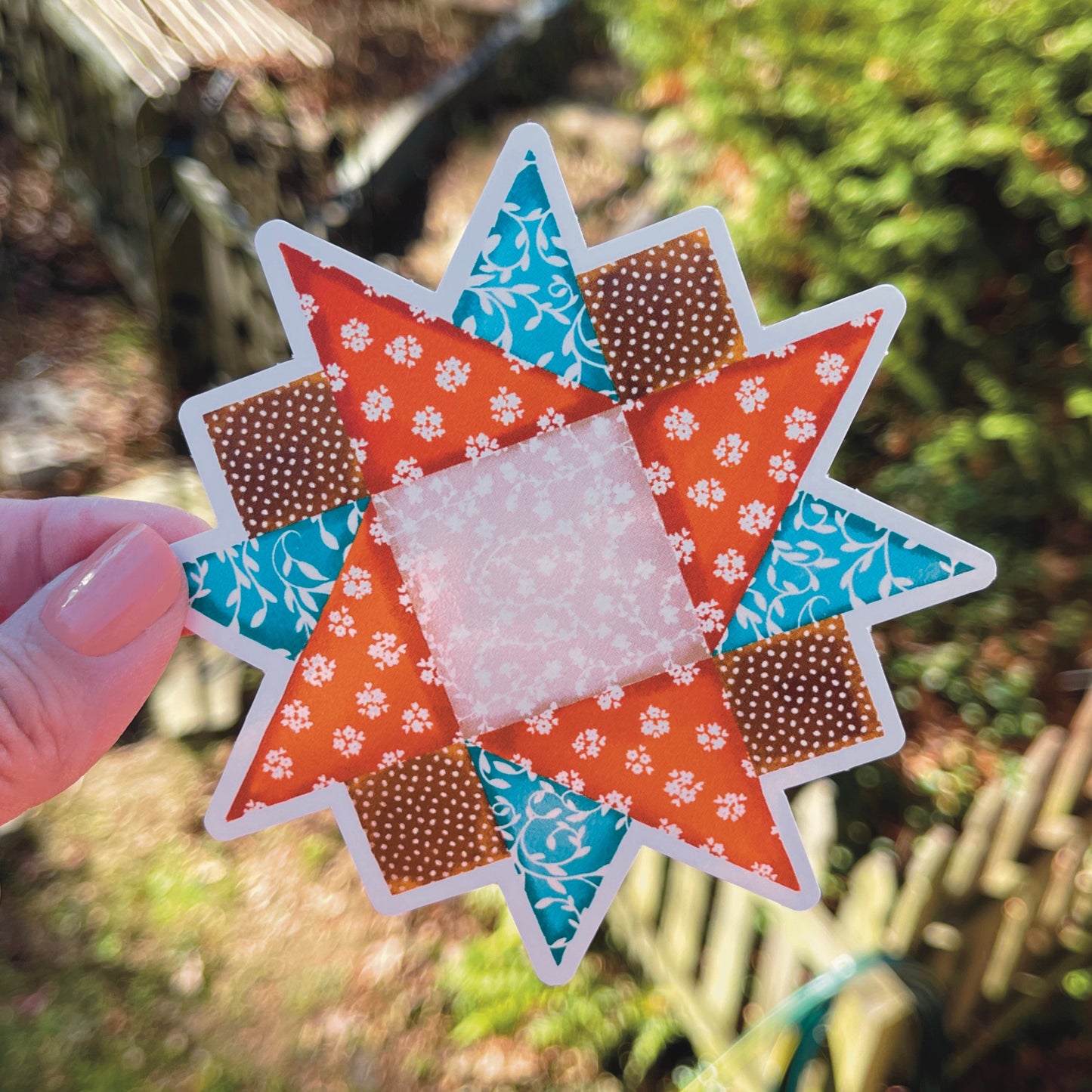 Medium orange, blue and brown quilt block sticker held over grassy background.