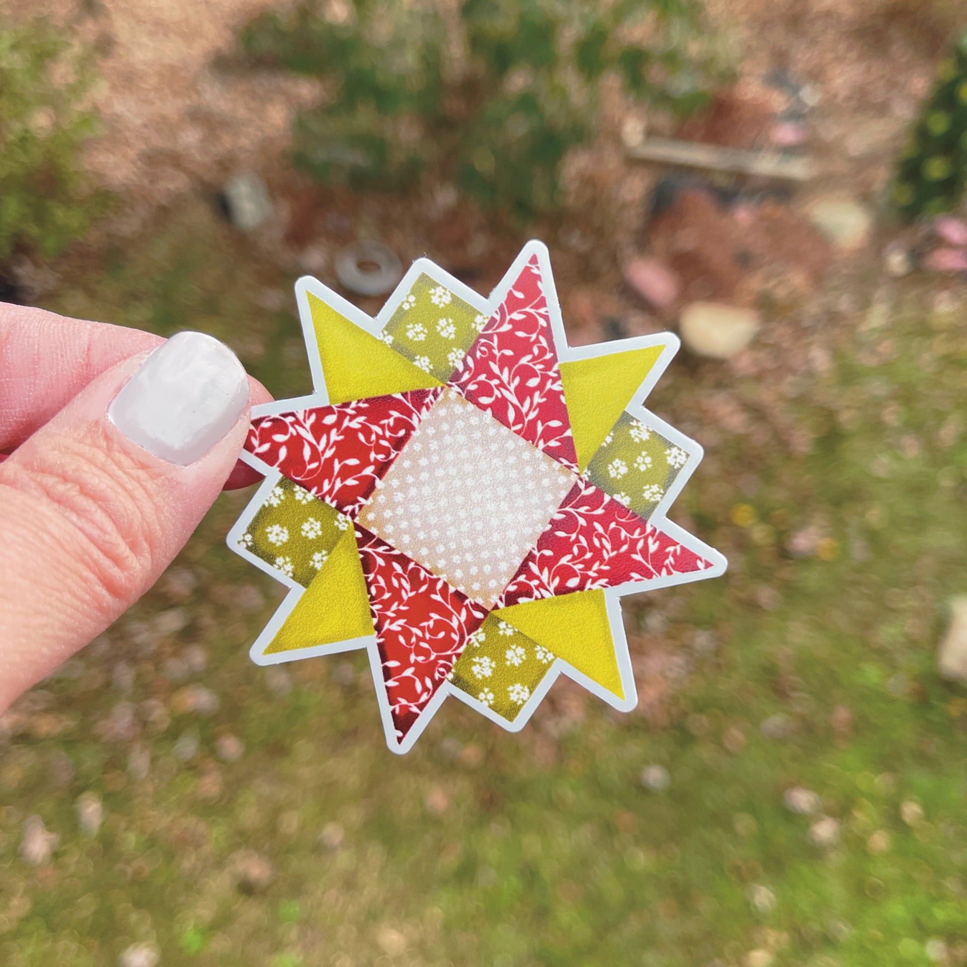 Red and green quilt block sticker held over grassy background.