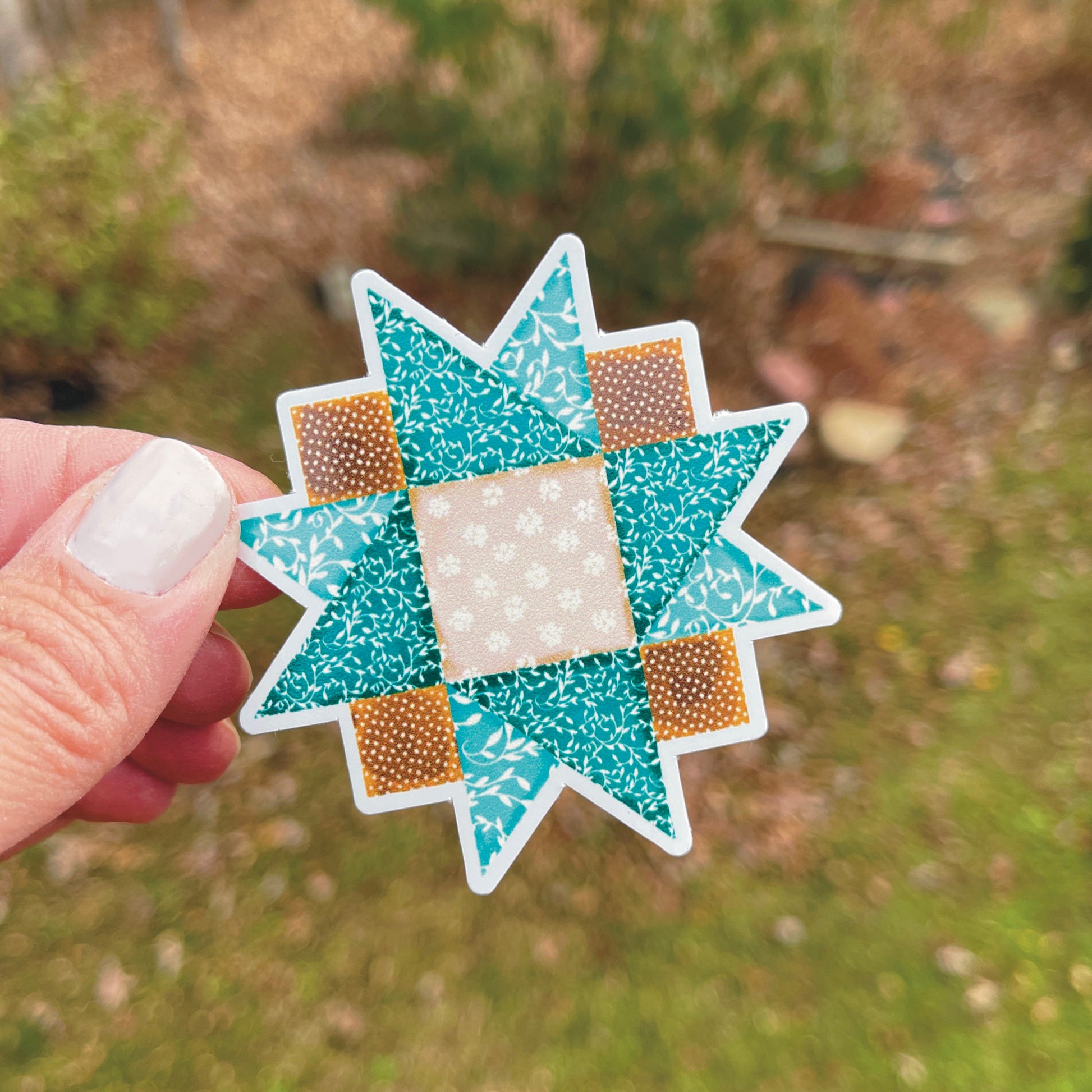 Small brown and blue quilt block sticker held over grassy background. 