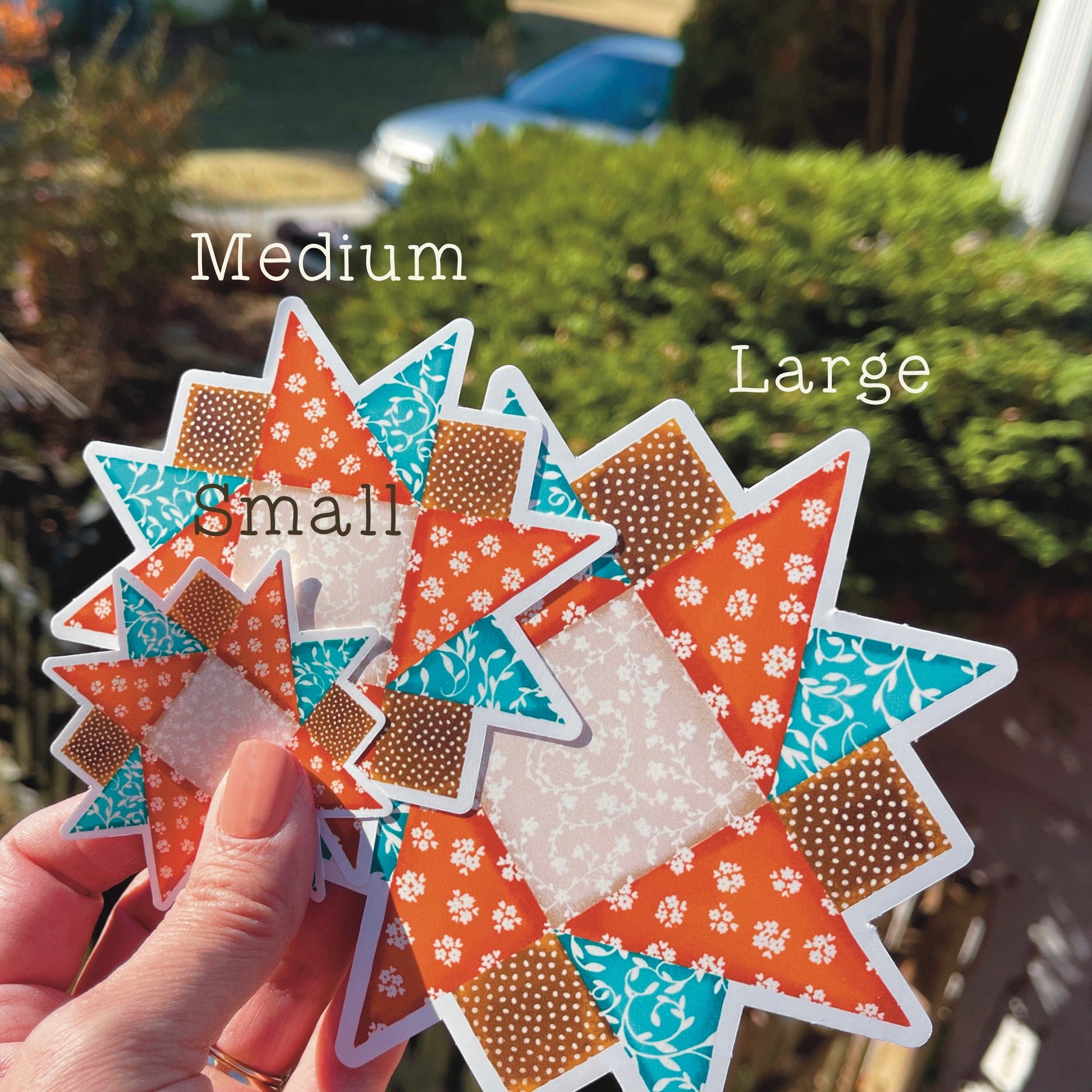 Three sizes of orange, blue and brown quilt block sticker held over grassy background.