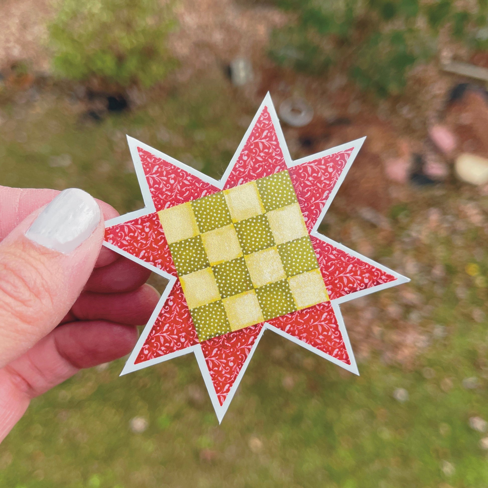 Red and green quilt block sticker held over grassy background.