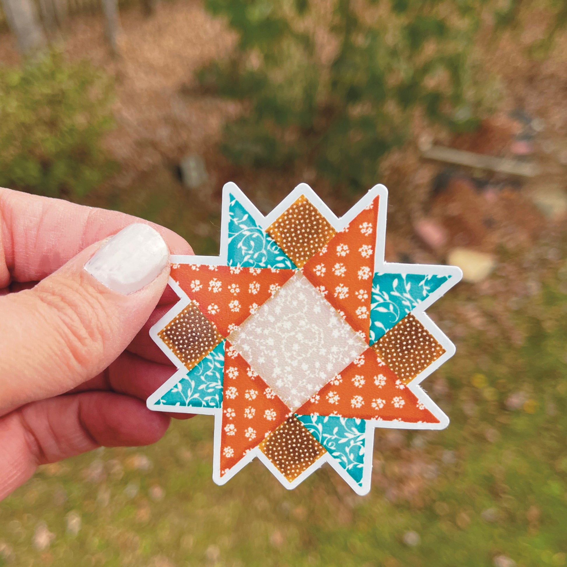 Small brown and blue quilt block sticker held over grassy background. 