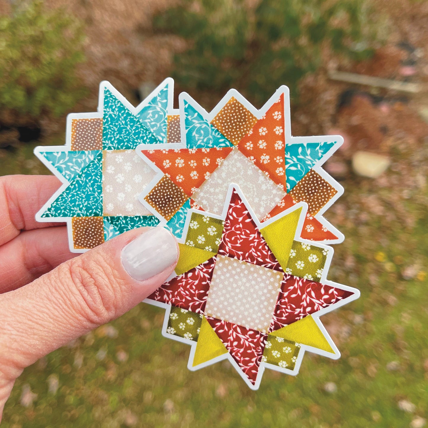 Set of three quilt block stickers blues, reds and orange held over a grassy background.