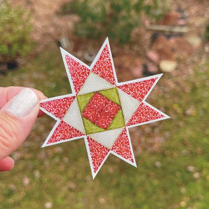 Red, Green and Tan Quilt block sticker held over grassy outside background. 