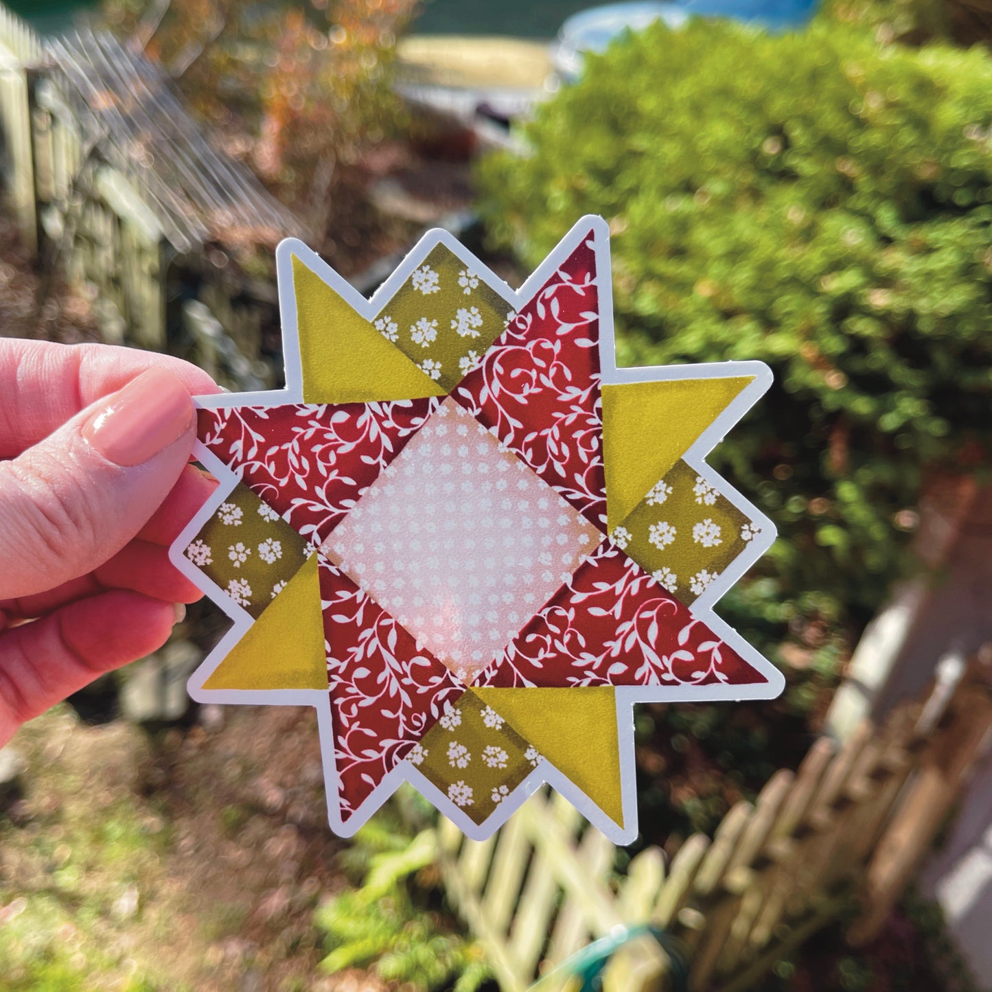 Medium red, green and tan quilt block sticker held over grassy background.