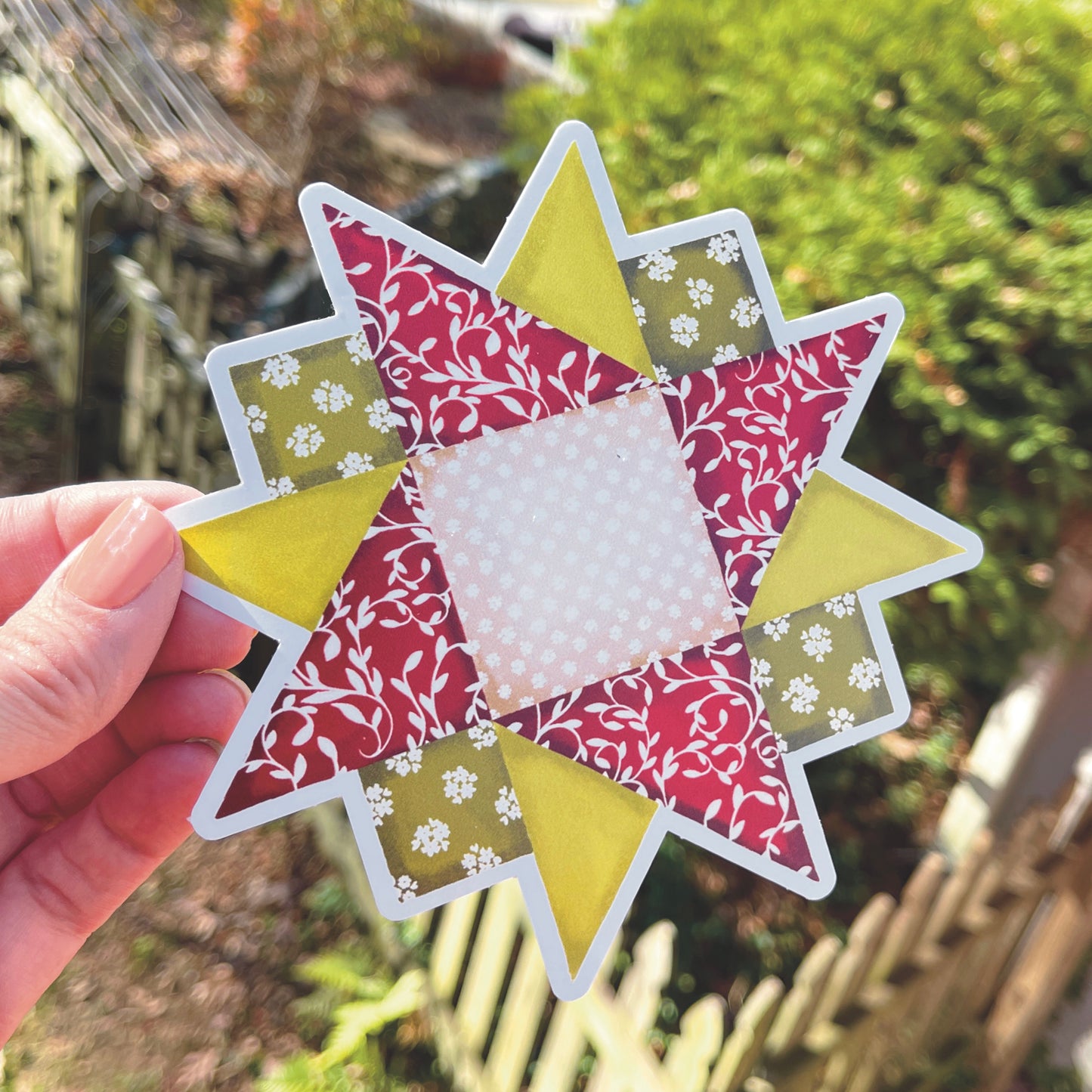 Large red, green and tan quilt block sticker held over grassy background.