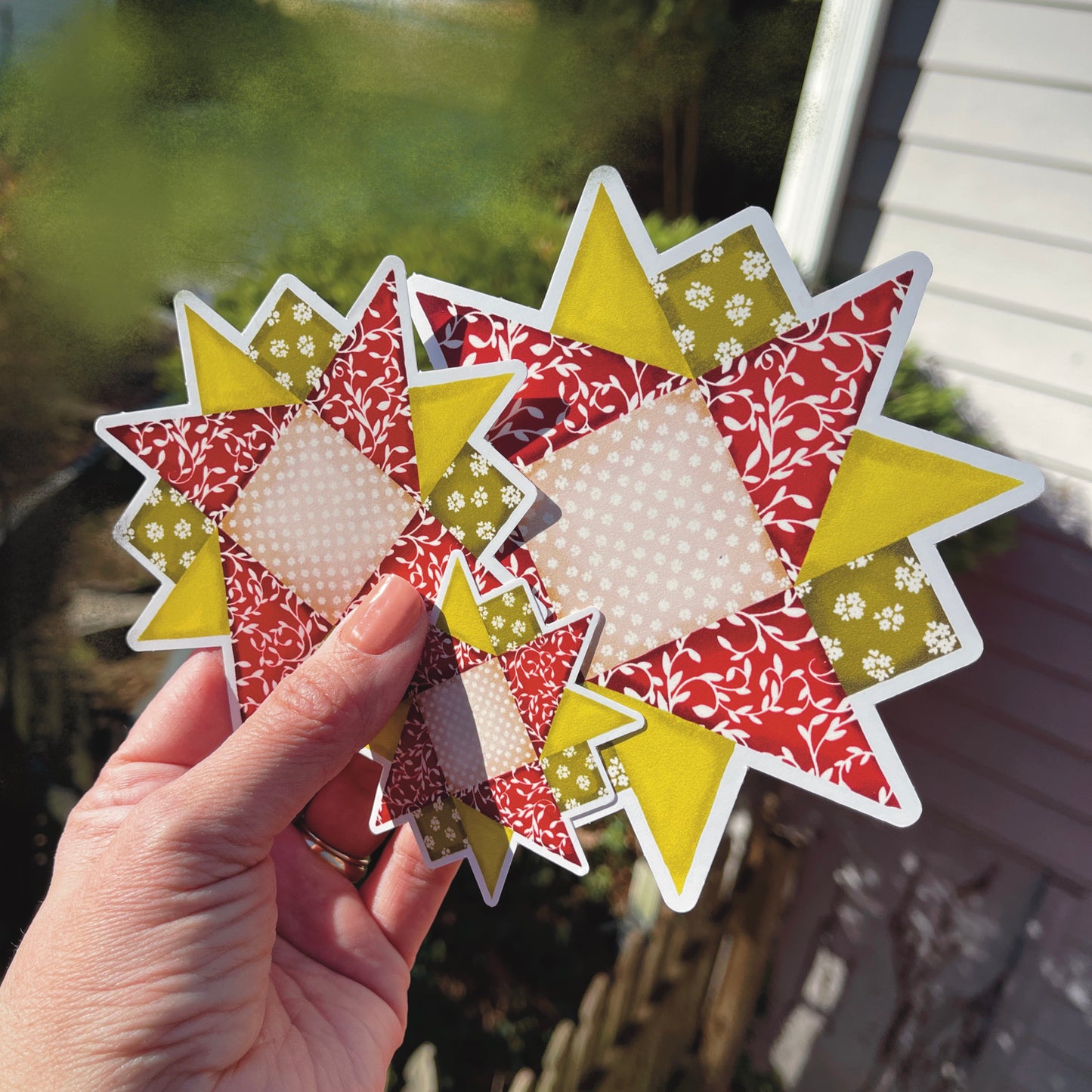 Three sizes of red, green and tan quilt block sticker held over grassy background.