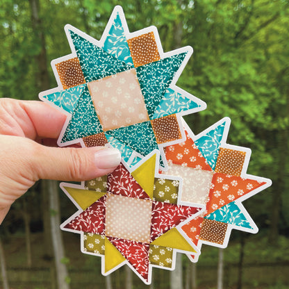 Set of three medium quilt block stickers blues, reds and orange held over a grassy background.