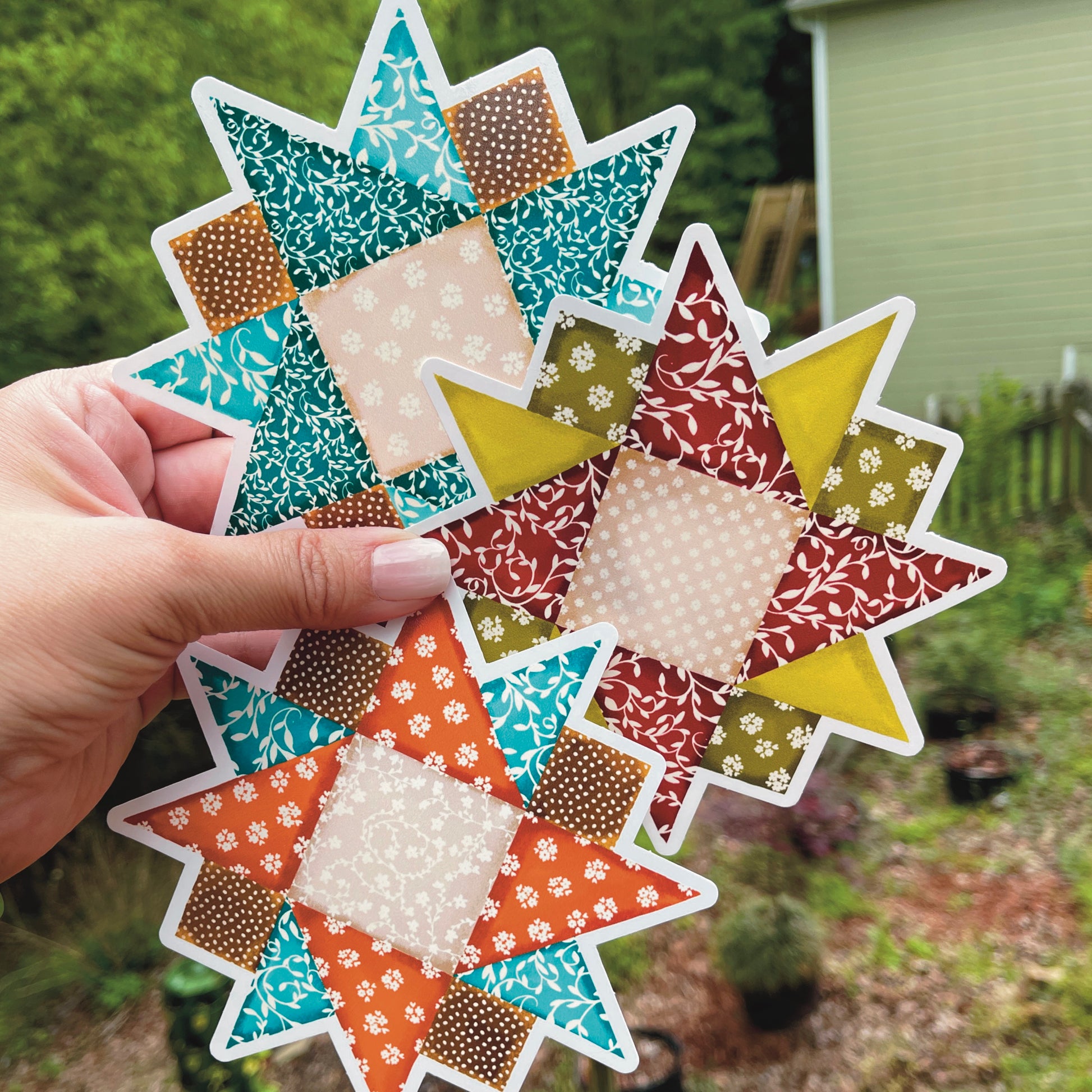 Set of three large quilt block stickers blues, reds and orange held over a grassy background.